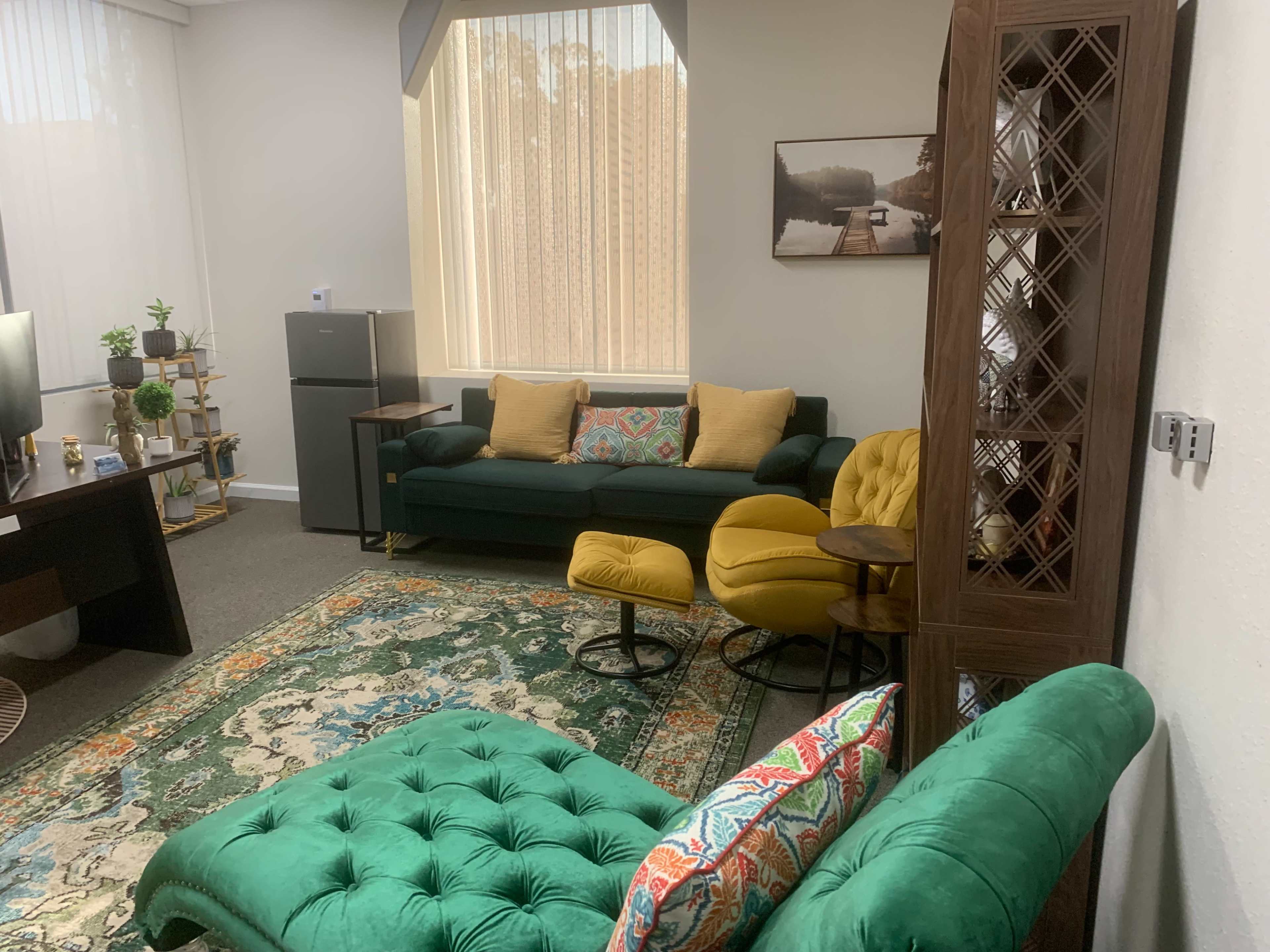 The image shows a cozy office space with a green sofa, yellow chairs, a wooden bookshelf, and a decorative rug.