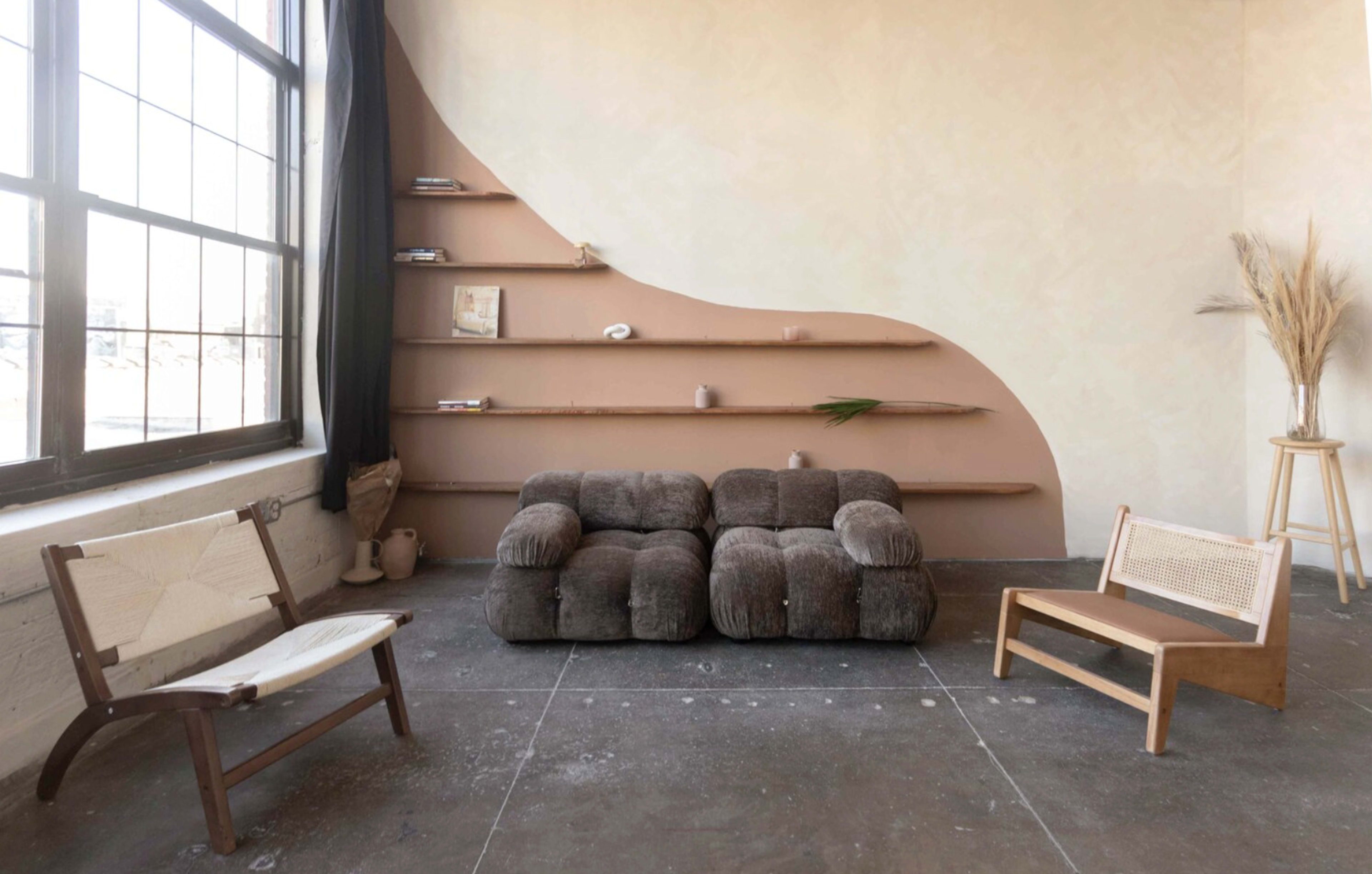 The image shows a minimalistic interior with a brown, tufted sofa, a wooden shelf against a curving wall, and two types of seating arrangements, all set in a spacious room with large windows.