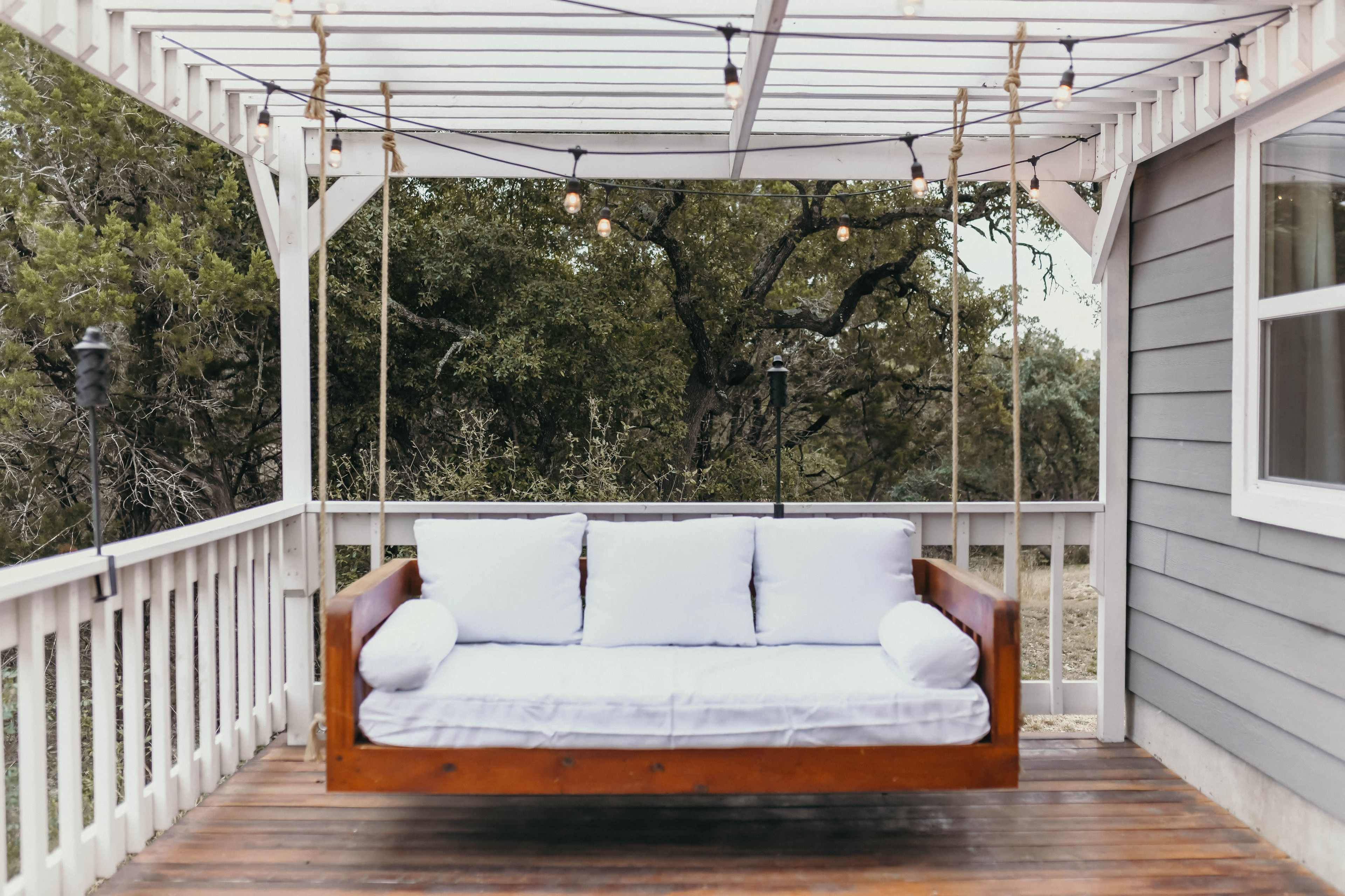 A wooden swing bed with white cushions hangs under a covered patio surrounded by trees.