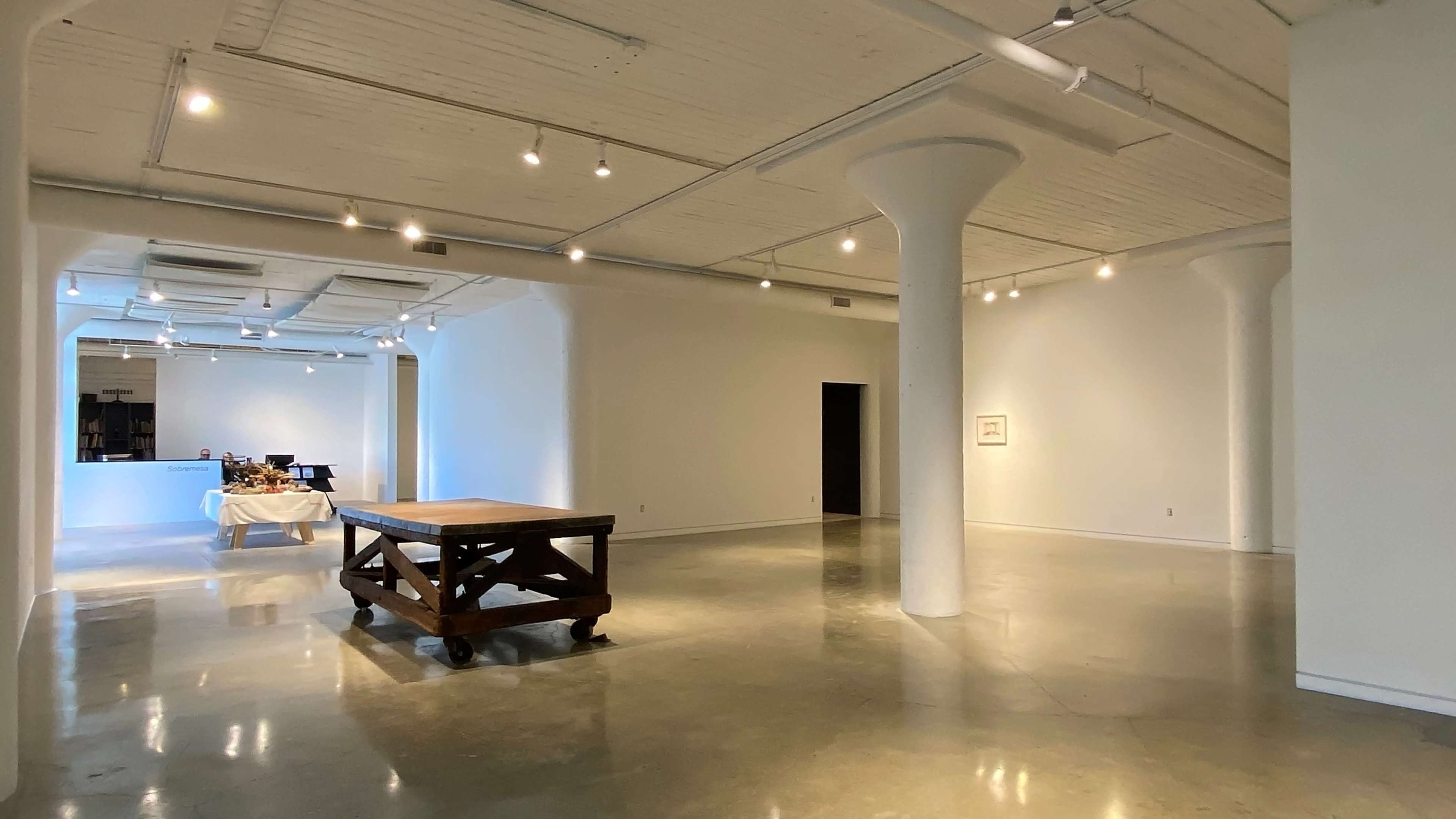 A spacious, well-lit interior of a modern gallery with polished concrete floors, featuring a large wooden table in the center and white walls.
