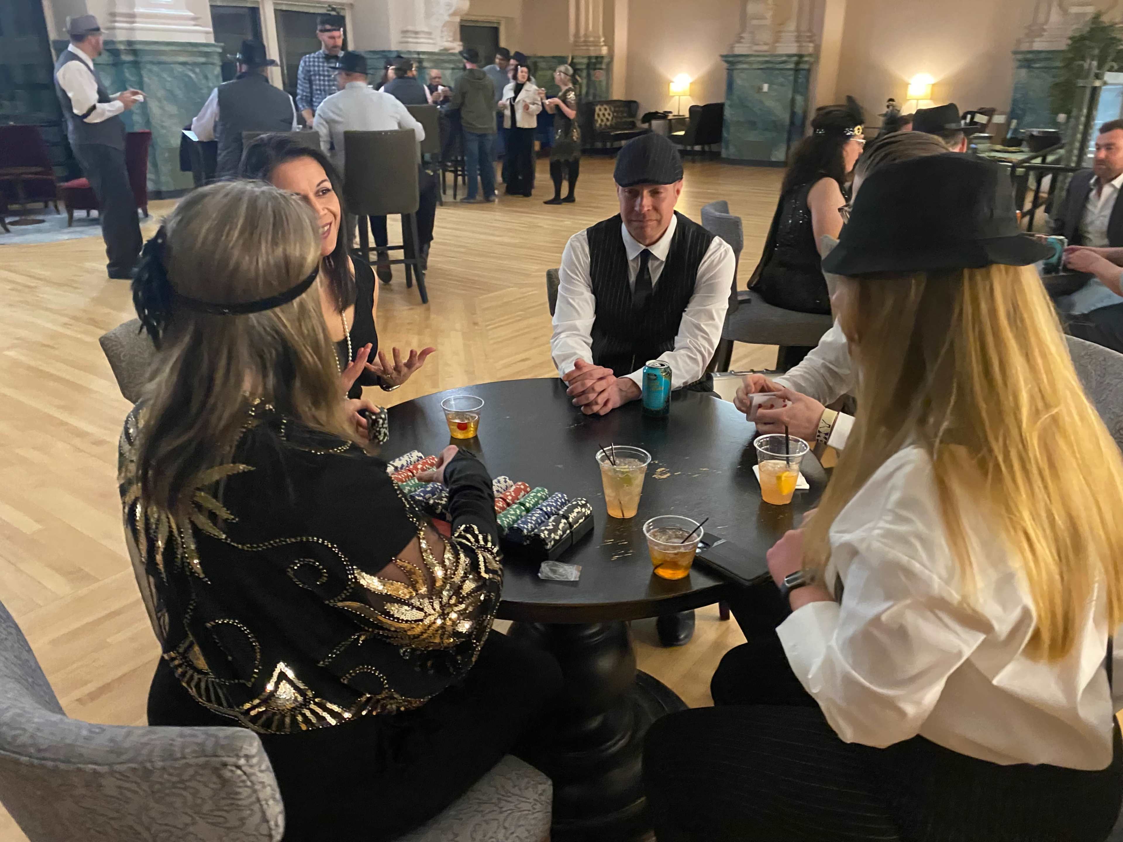 A group of people dressed in vintage attire sits around a table with drinks and poker chips in a spacious room with high ceilings.