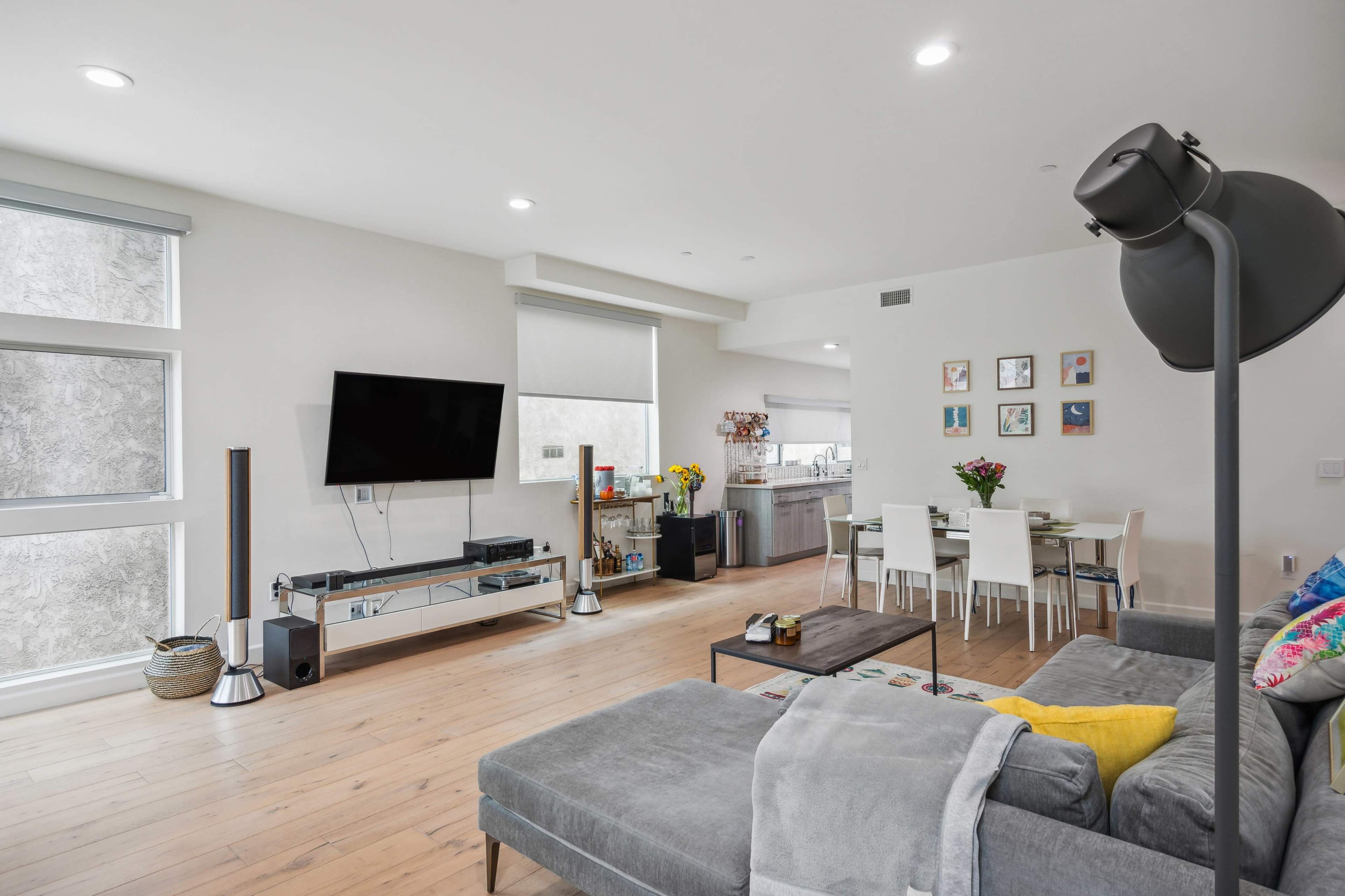 The image shows a modern living room with a gray sectional sofa, a large flat-screen TV mounted on the wall, and a dining area with white chairs and a table.