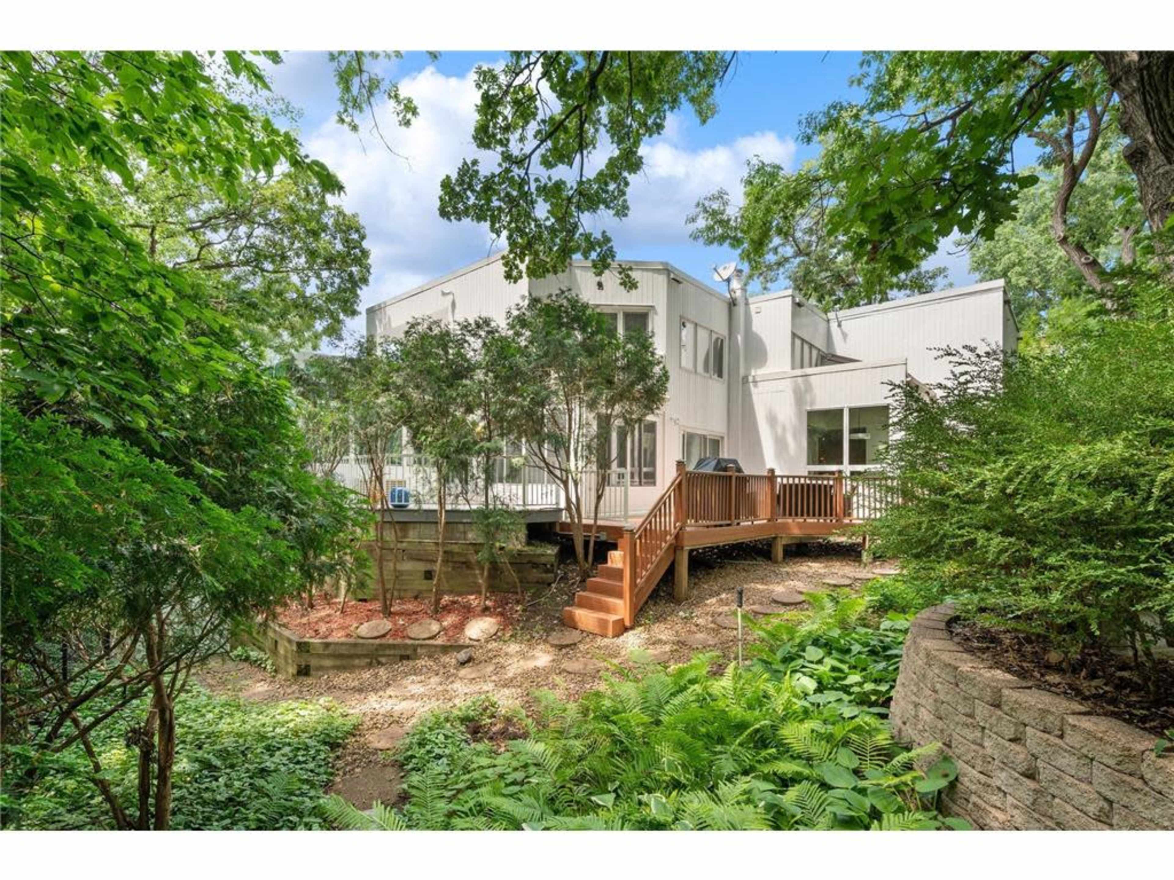 The image shows a modern house partially surrounded by greenery, featuring a deck that extends into a landscaped garden area.
