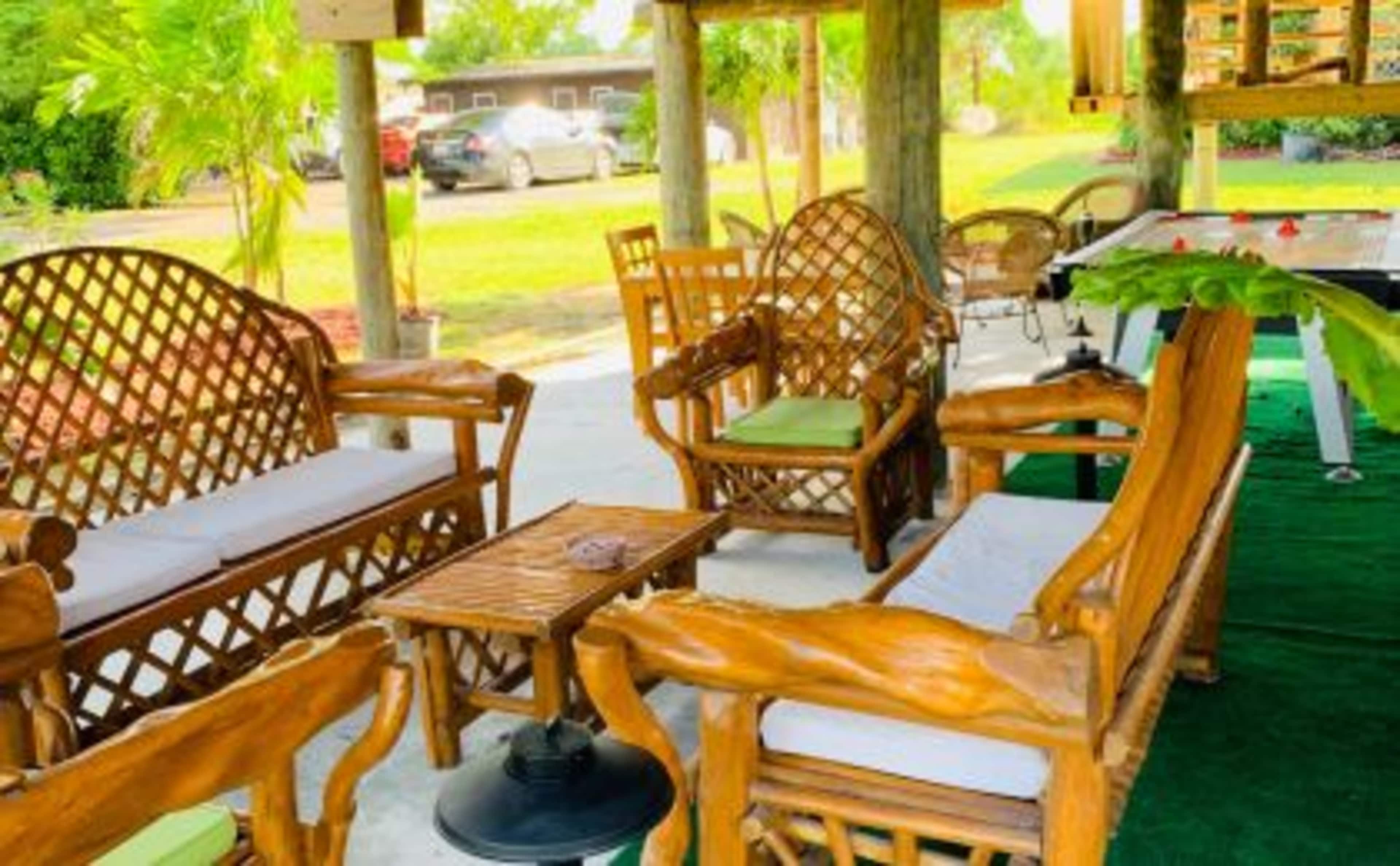 The image shows a shaded outdoor seating area featuring wooden furniture, including chairs and a small table, surrounded by greenery and a foosball table in the background.
