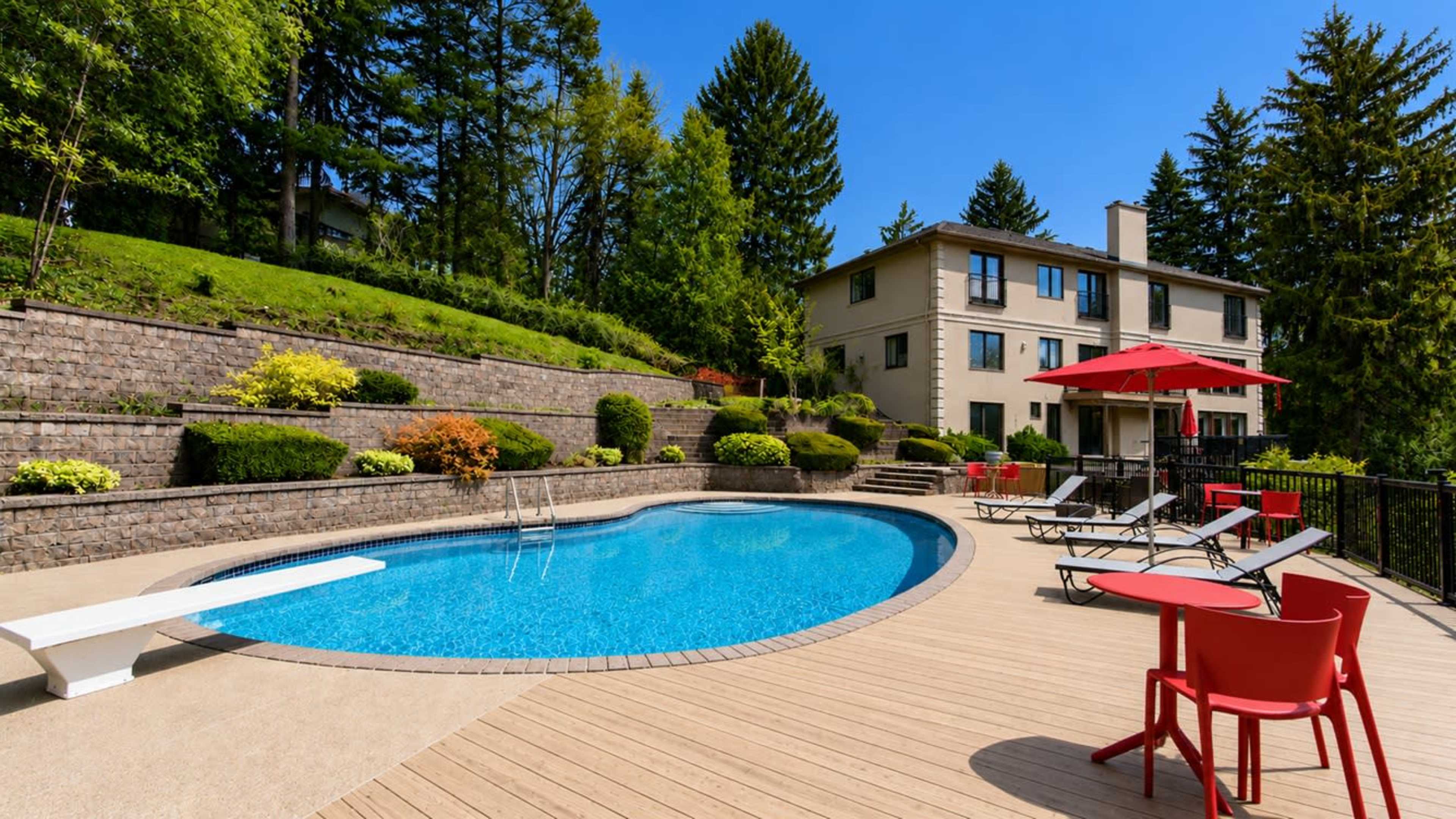 The image depicts a landscaped backyard with a blue swimming pool, lounge chairs, a diving board, and a two-story house in the background.