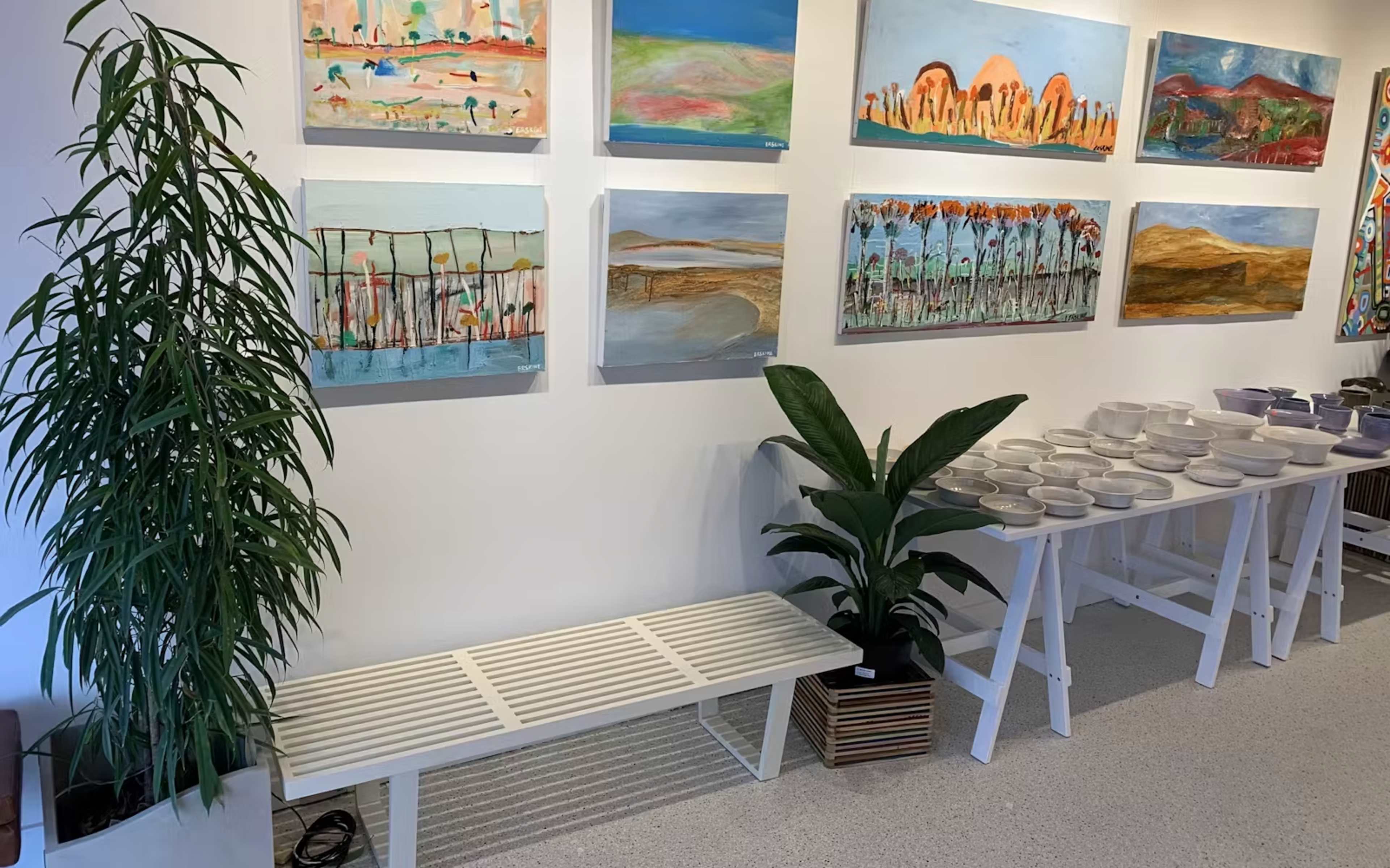 A gallery wall adorned with several colorful paintings, alongside a plant and a display of white dishware on a white table.