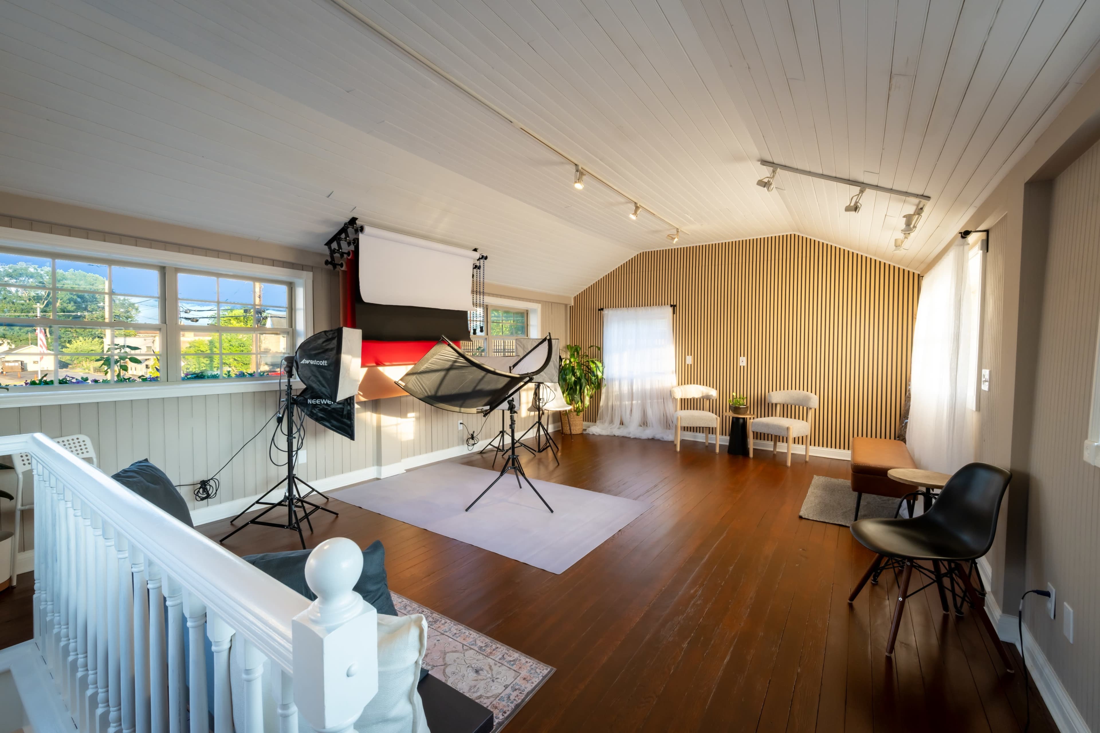 The image shows a spacious indoor studio with wooden floors, large windows, and photography equipment set up, including softbox lights and a backdrop.