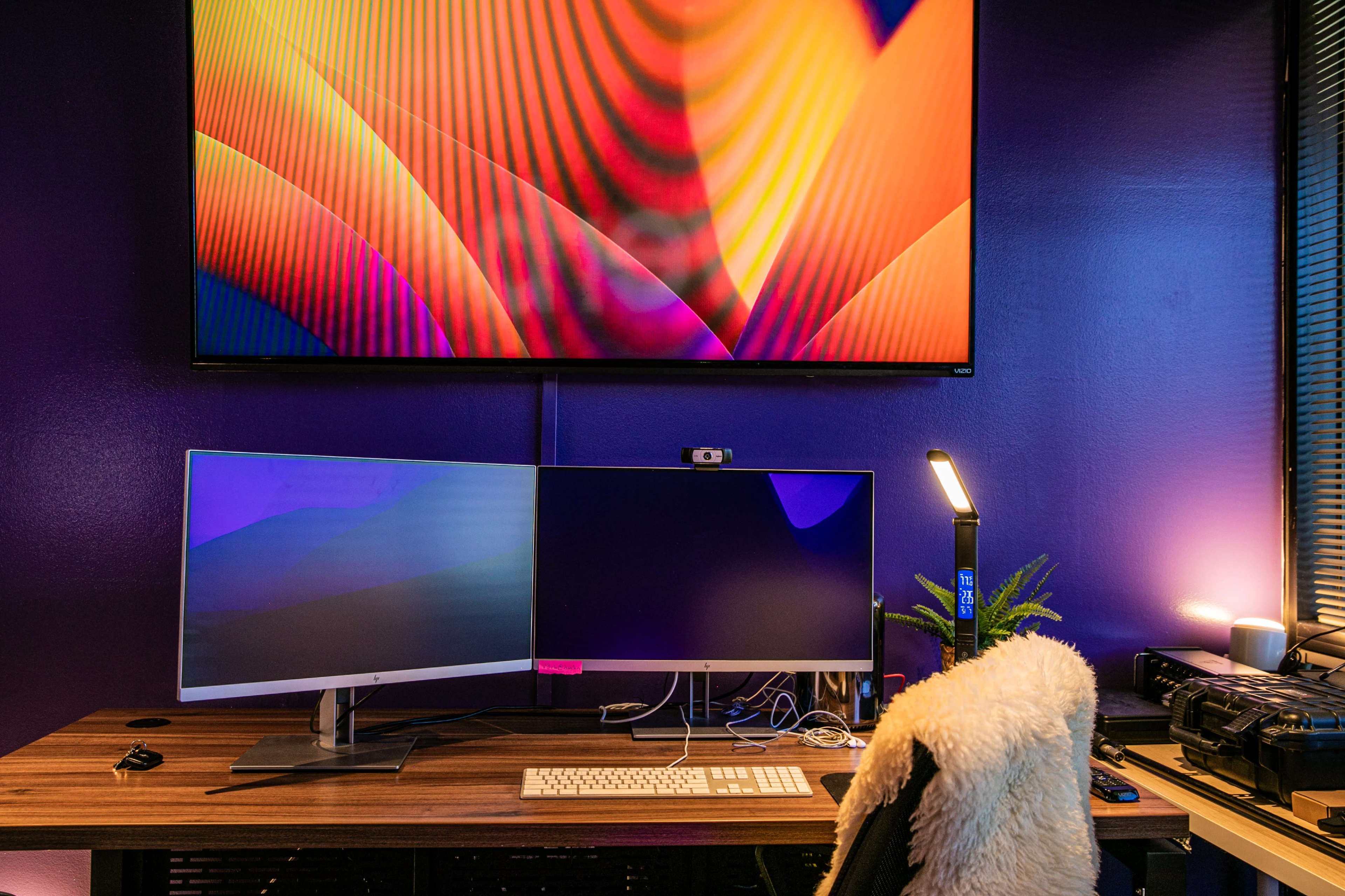 A workspace with a dual-monitor setup on a wooden desk, accompanied by a large wall-mounted screen displaying colorful graphics and a desk lamp.