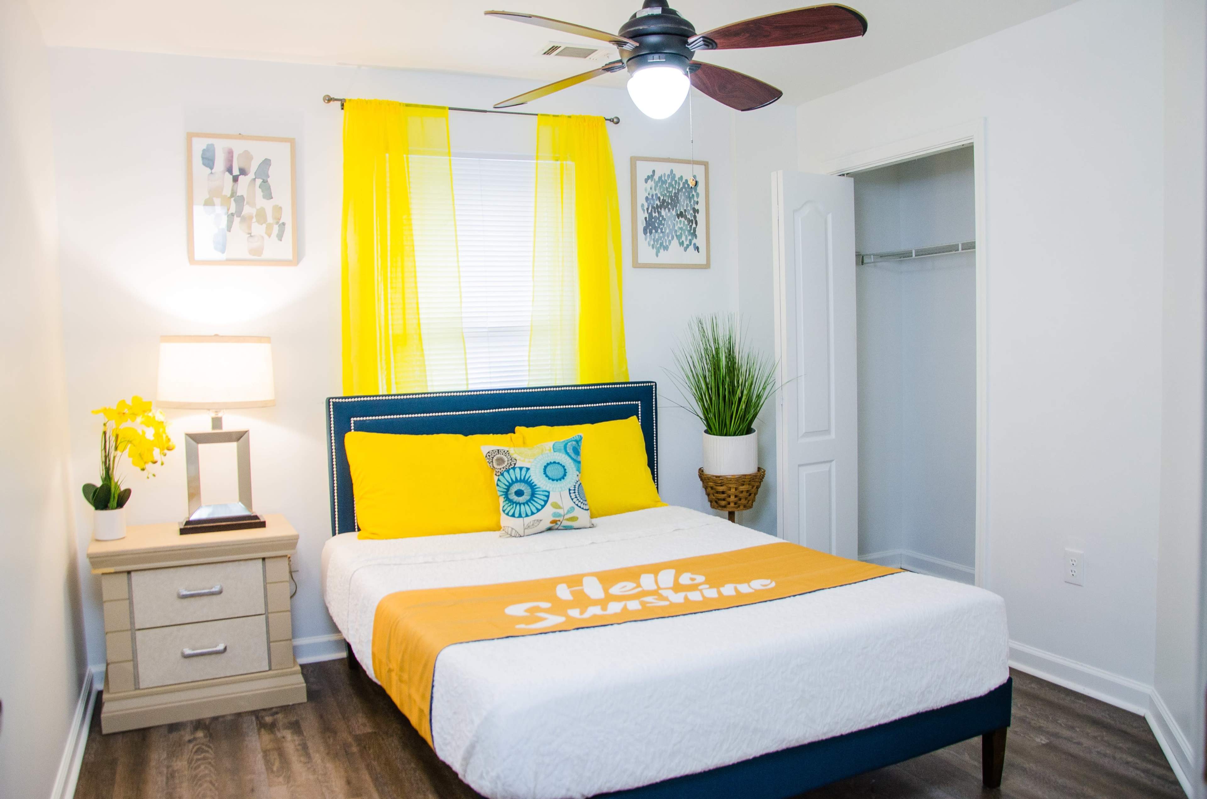 A well-lit bedroom features a bed with a yellow blanket, a nightstand with a lamp, and bright curtains by the window.
