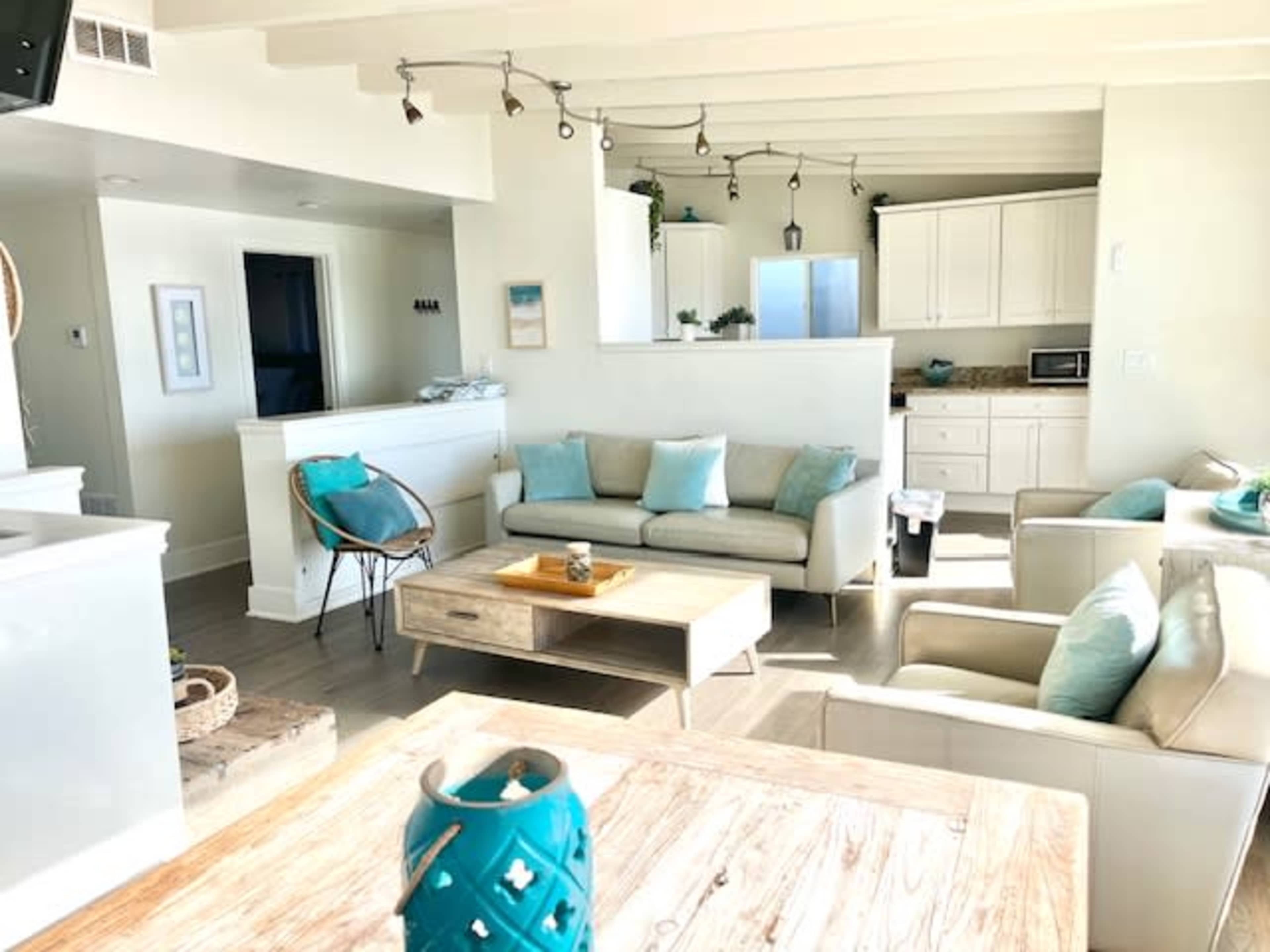 The image shows a bright living room with light-colored furniture, a wooden coffee table, and an open kitchen space in the background.