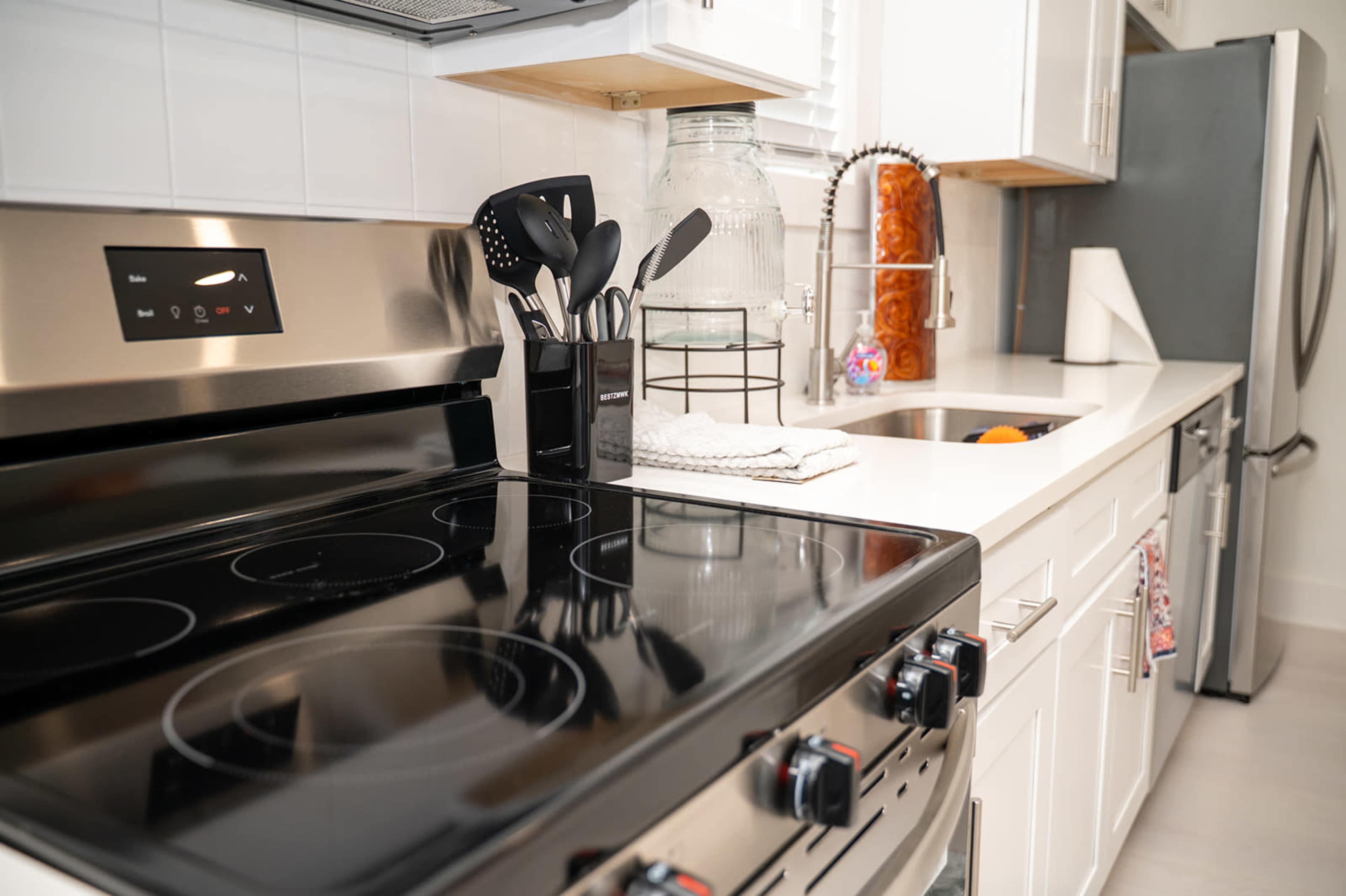 The image shows a modern kitchen with a black stove, stainless steel appliances, and a countertop with cooking utensils and a glass jar.