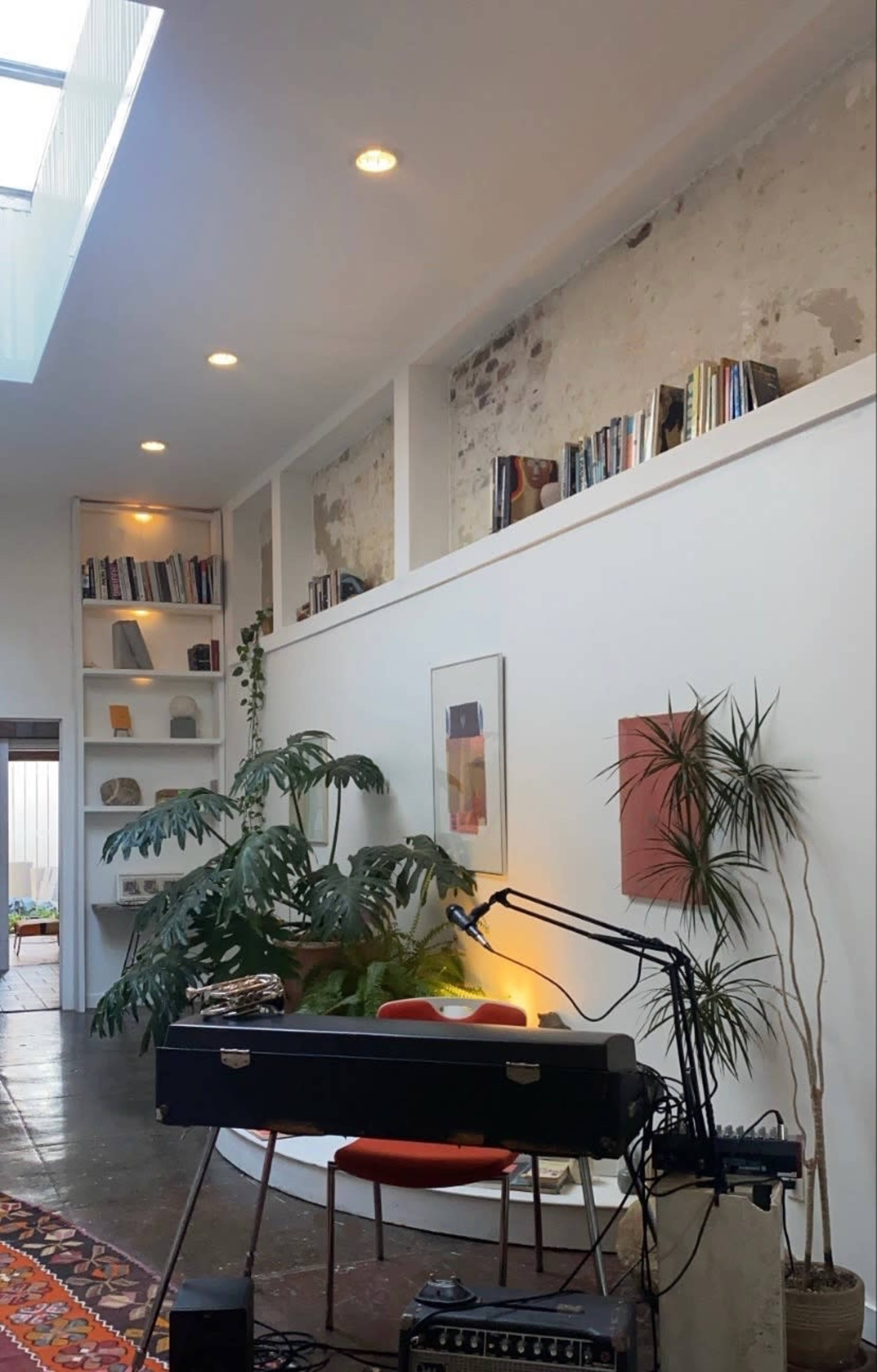 A spacious interior with a piano, a microphone stand, and a wall-mounted bookshelf, complemented by large plants and natural light from a skylight.