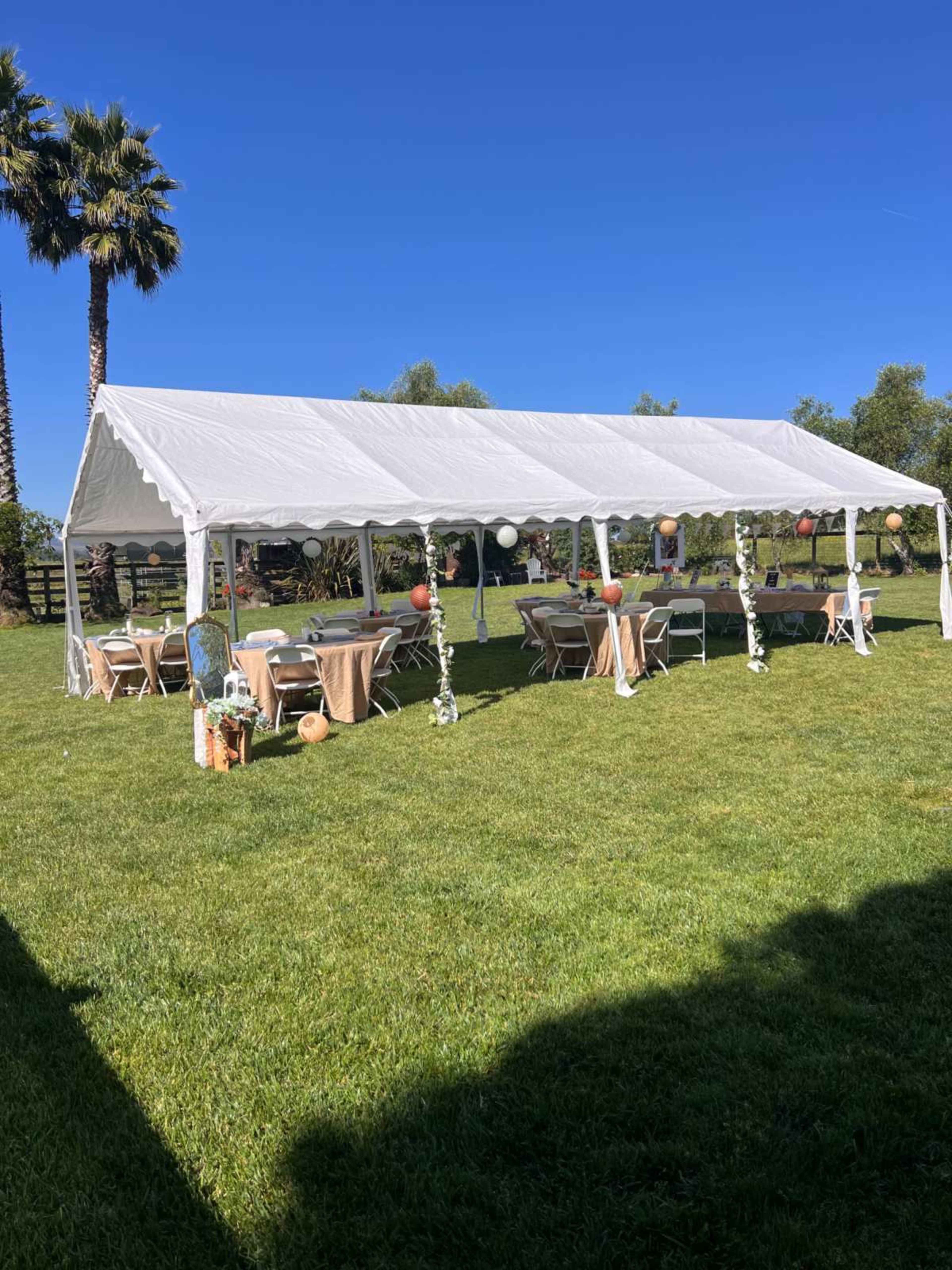 A large white tent is set up on a grassy area, surrounded by palm trees, with tables covered in beige cloth and decorative elements.