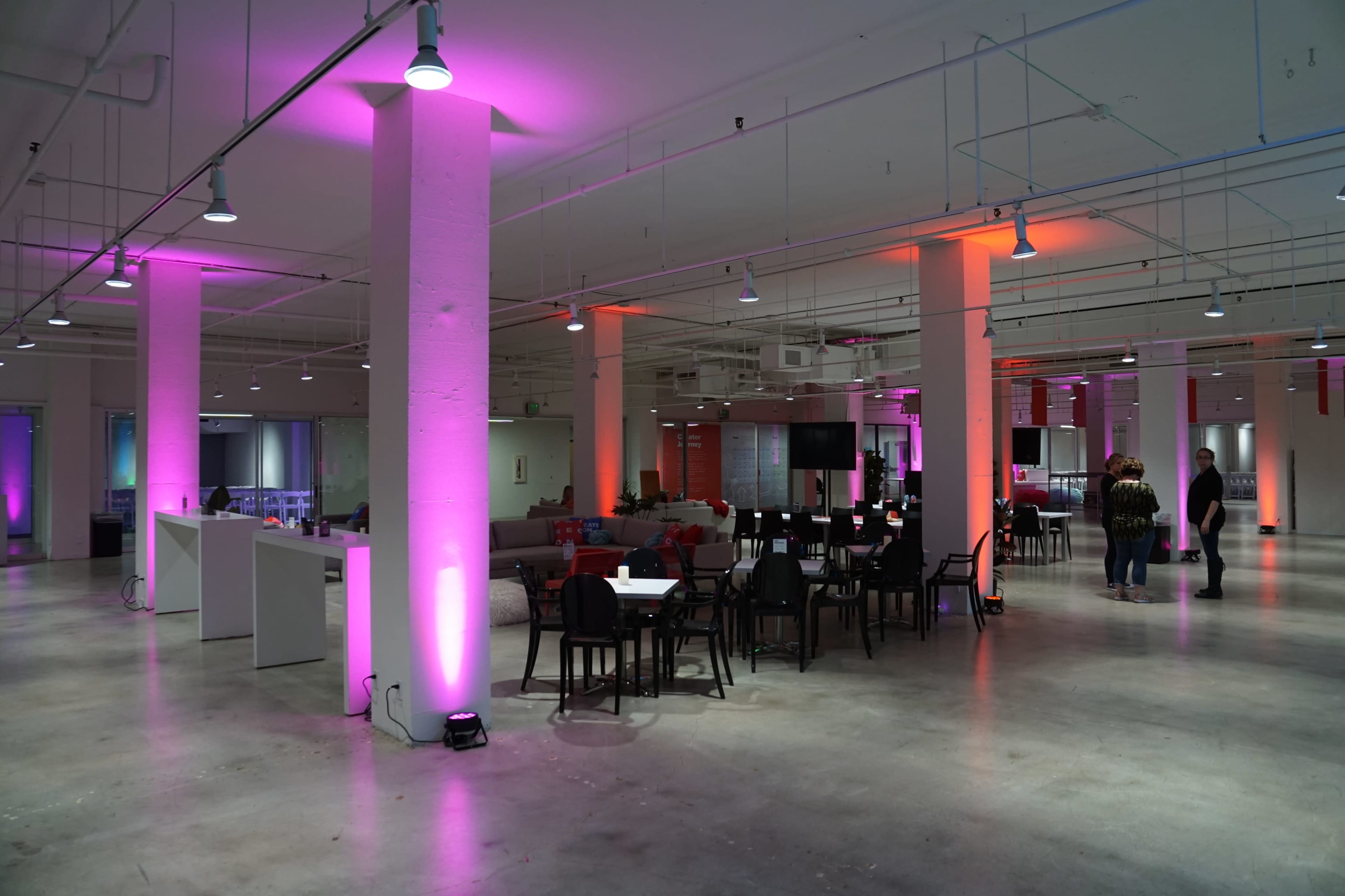 The image depicts a spacious, modern interior with pink and orange lighting, featuring tables and chairs arranged throughout the area.