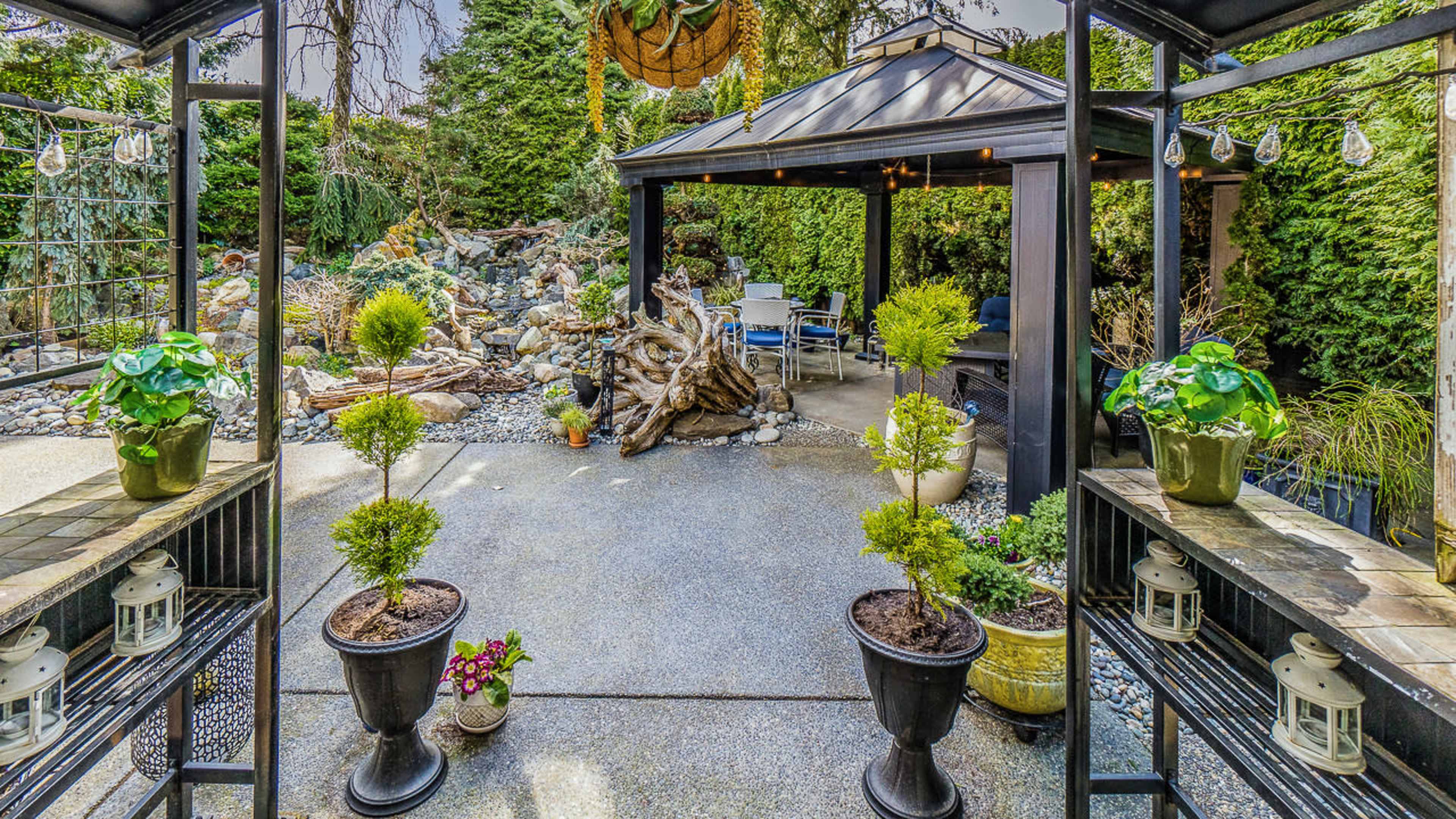 The image shows a landscaped outdoor space featuring a gazebo surrounded by greenery, decorative plants, and lanterns.