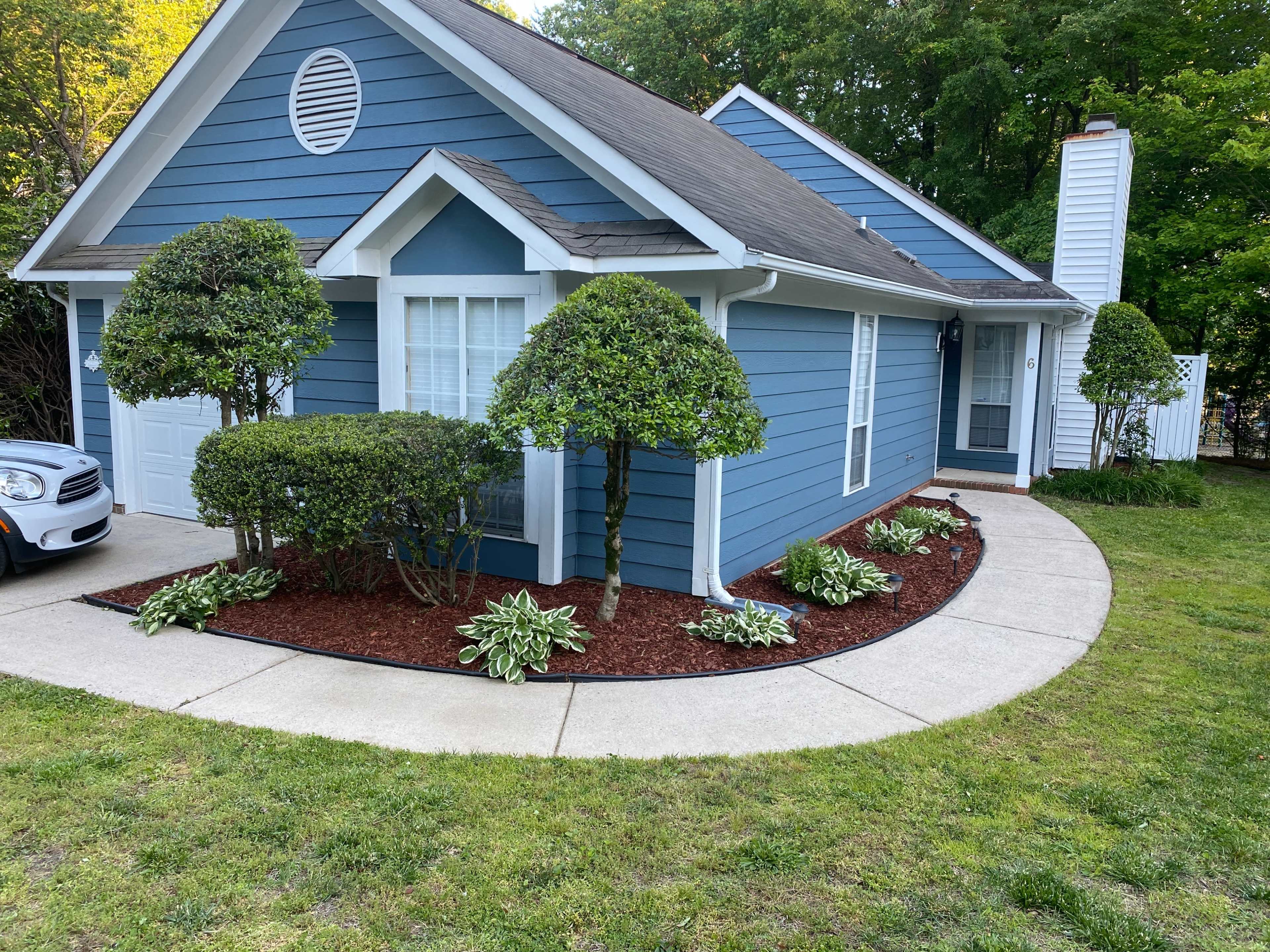 The image shows a blue house with a neatly manicured front yard featuring bushes and flower beds along a curved concrete walkway.