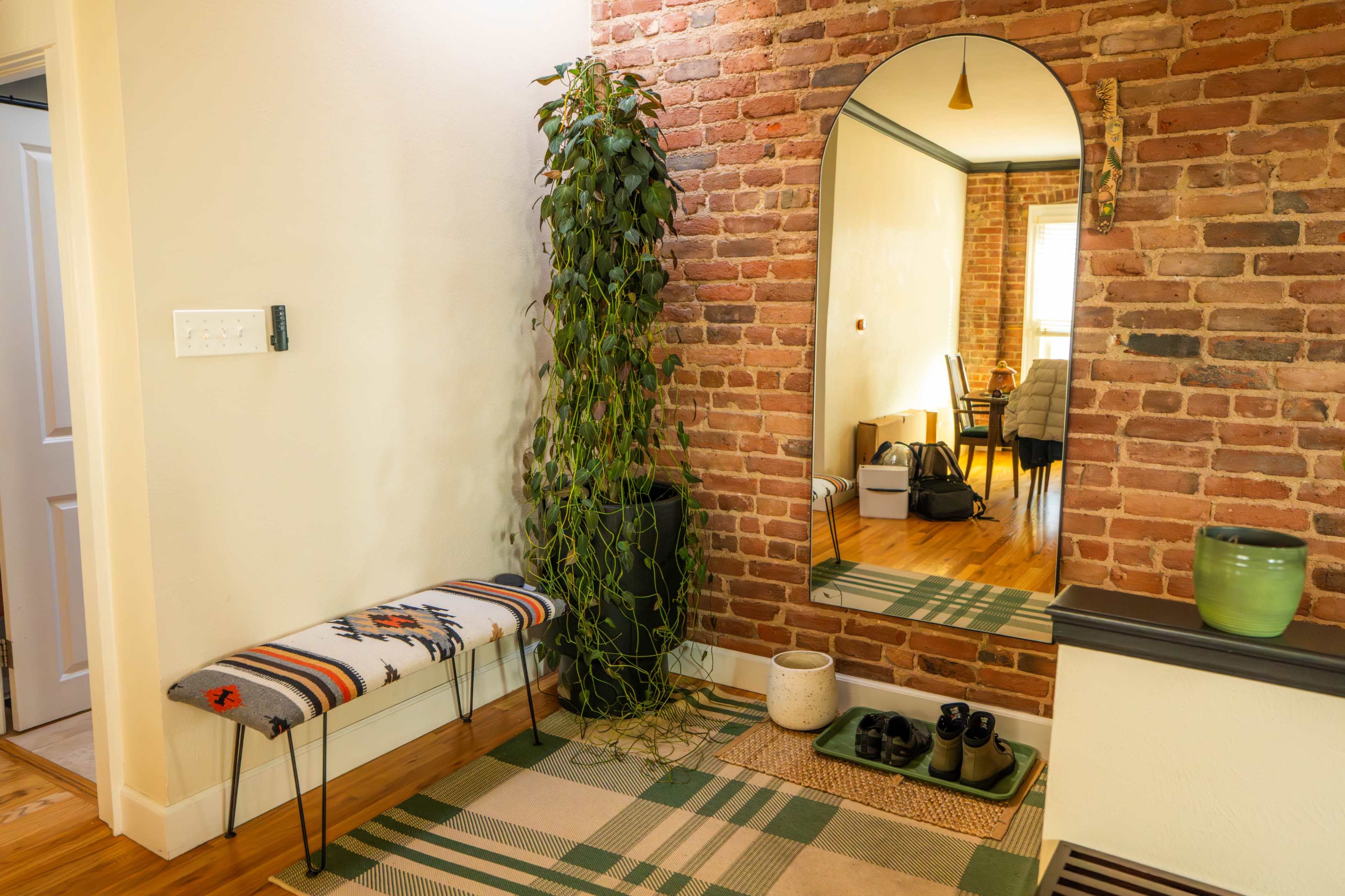 The image shows a small entryway with a brick wall, a mirror, a plant draping down from a tall pot, a bench, and a patterned rug.