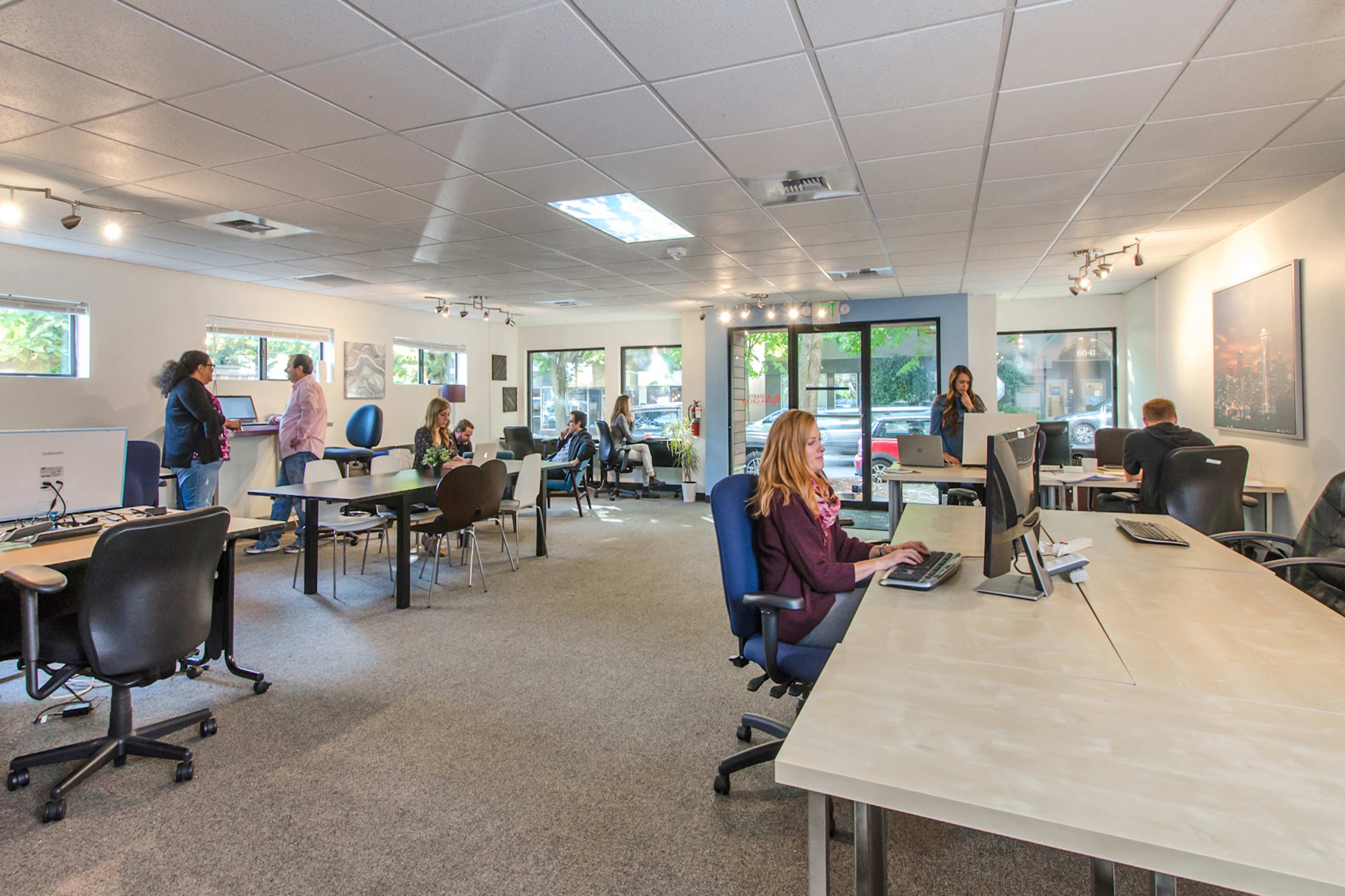The image shows an office space with several people working at desks and using computers in a well-lit environment.