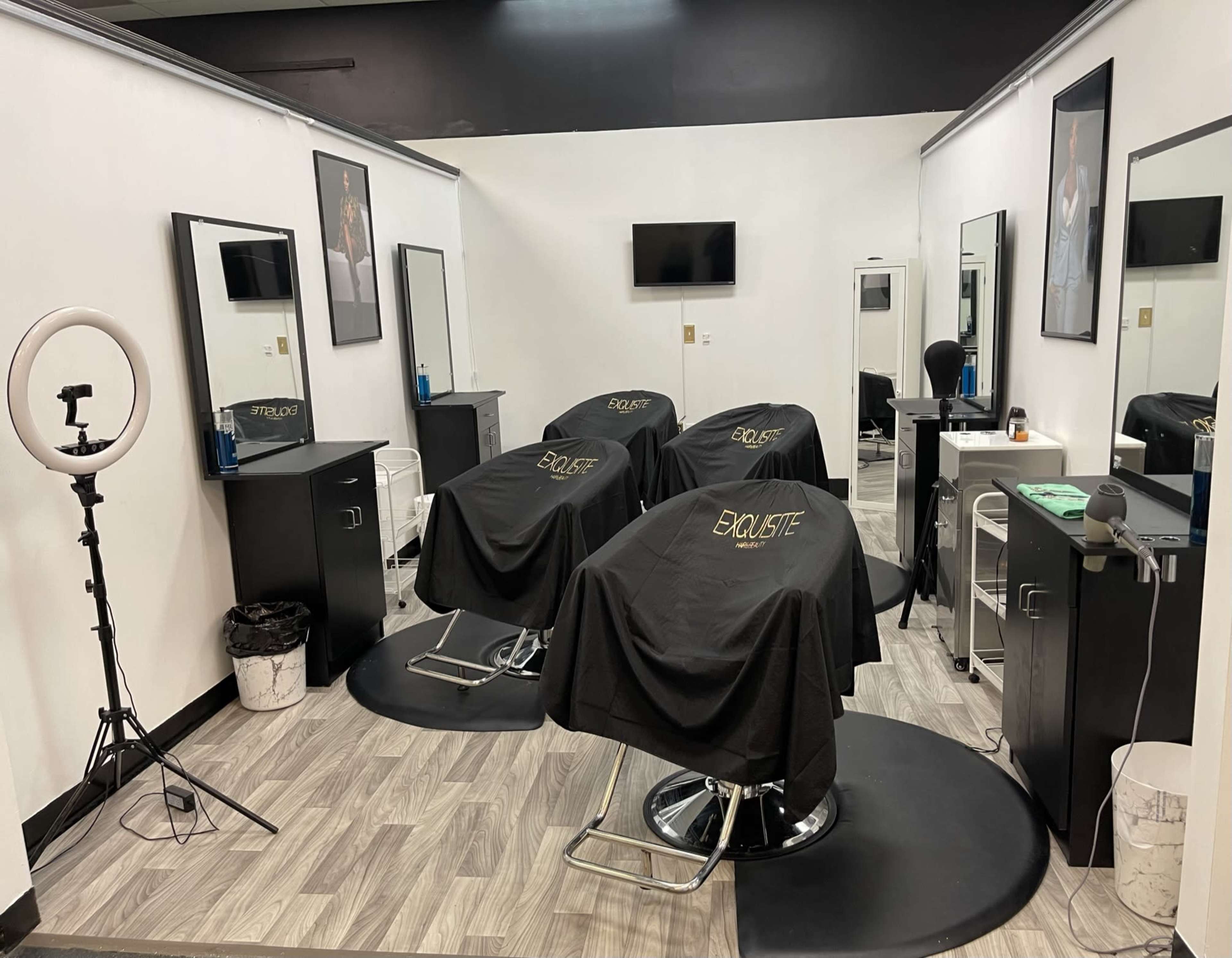 A modern hair salon with three styling chairs, each covered with a black cape, surrounded by mirrors and illuminated by a ring light.