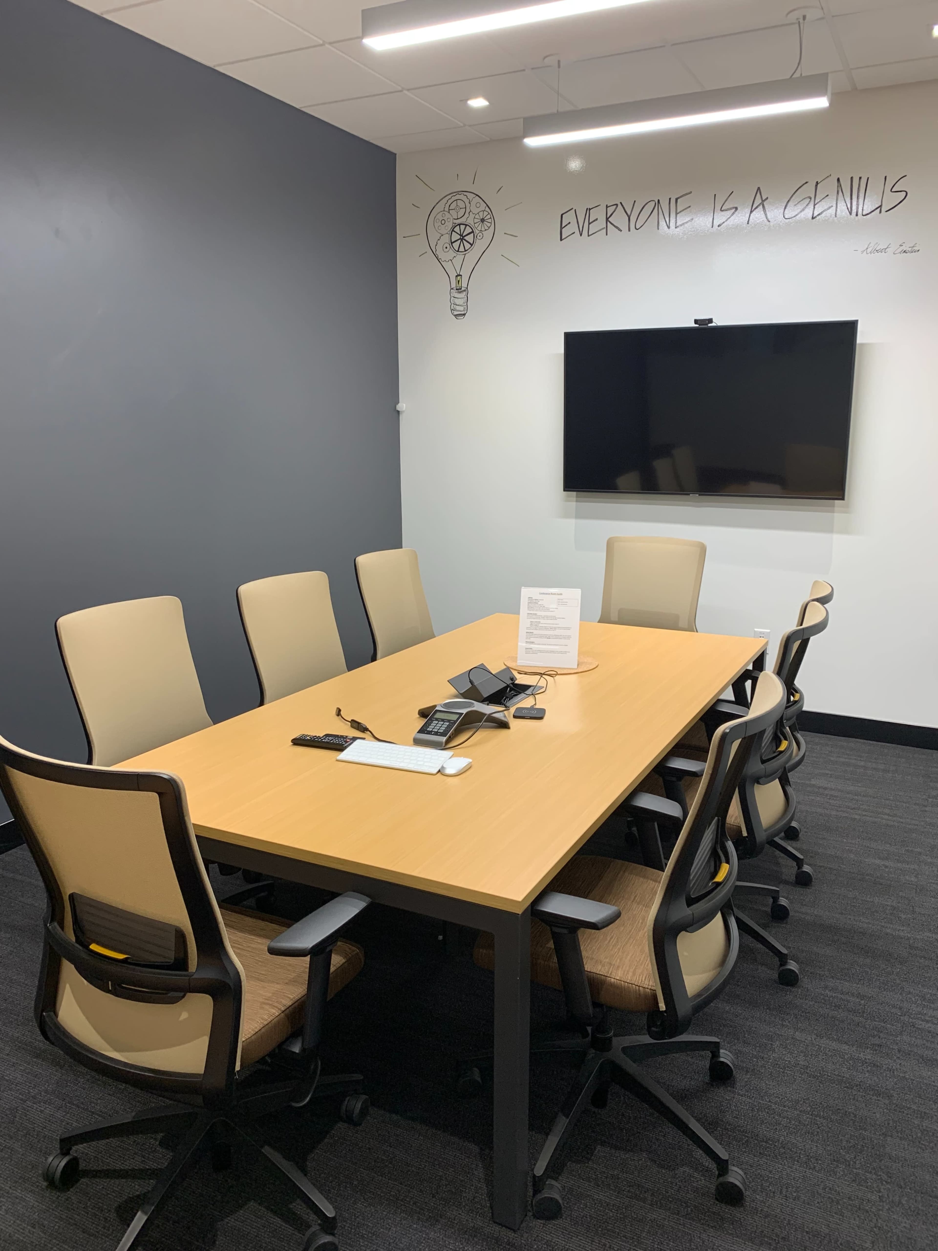 A conference room features a long wooden table surrounded by eight chairs, a wall-mounted television, and a whiteboard with a quote written above it.