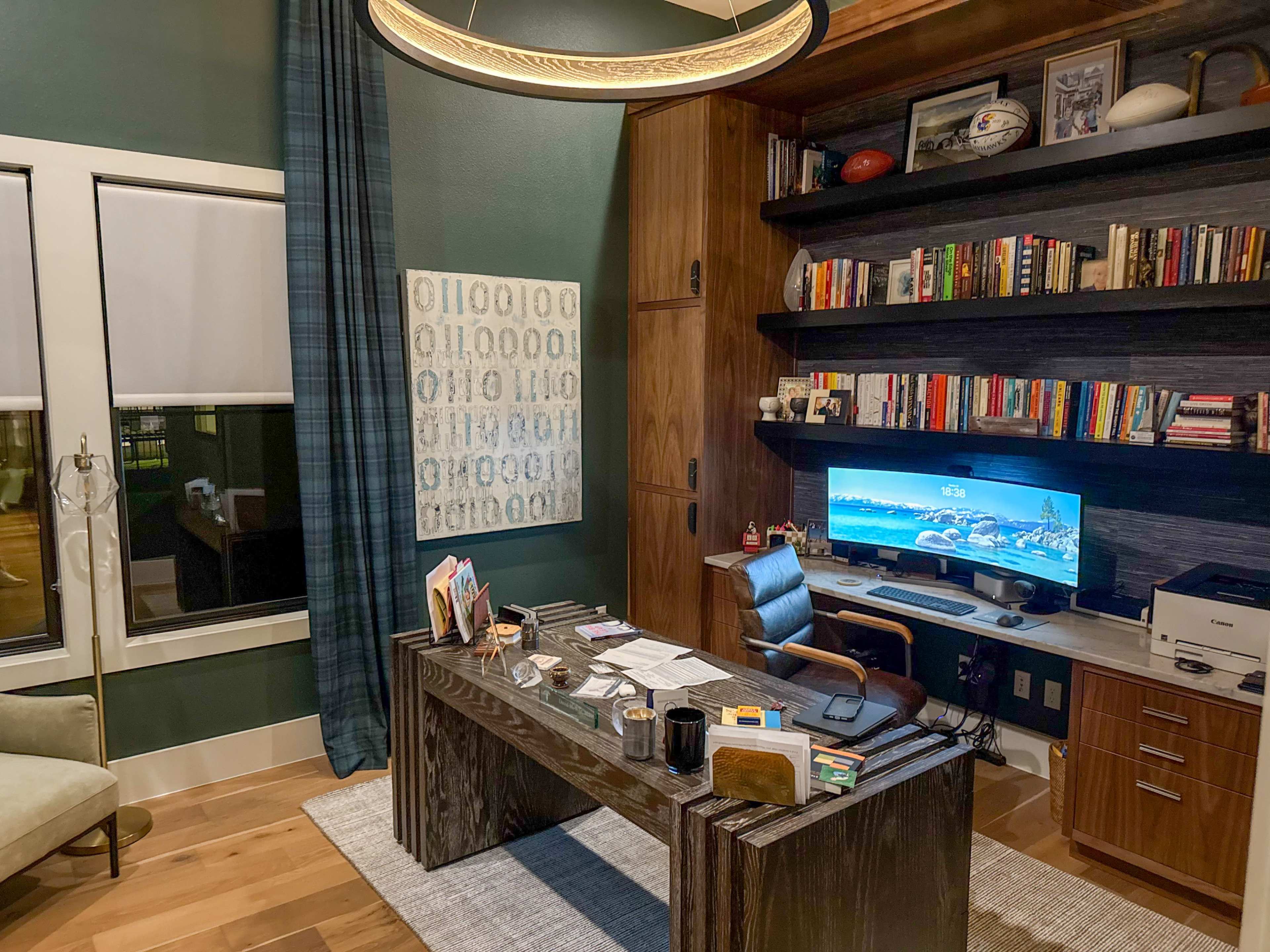 The image shows a modern home office featuring a large wooden desk, a comfortable chair, a wall-mounted TV, bookshelves filled with books, and a window with blackout curtains.