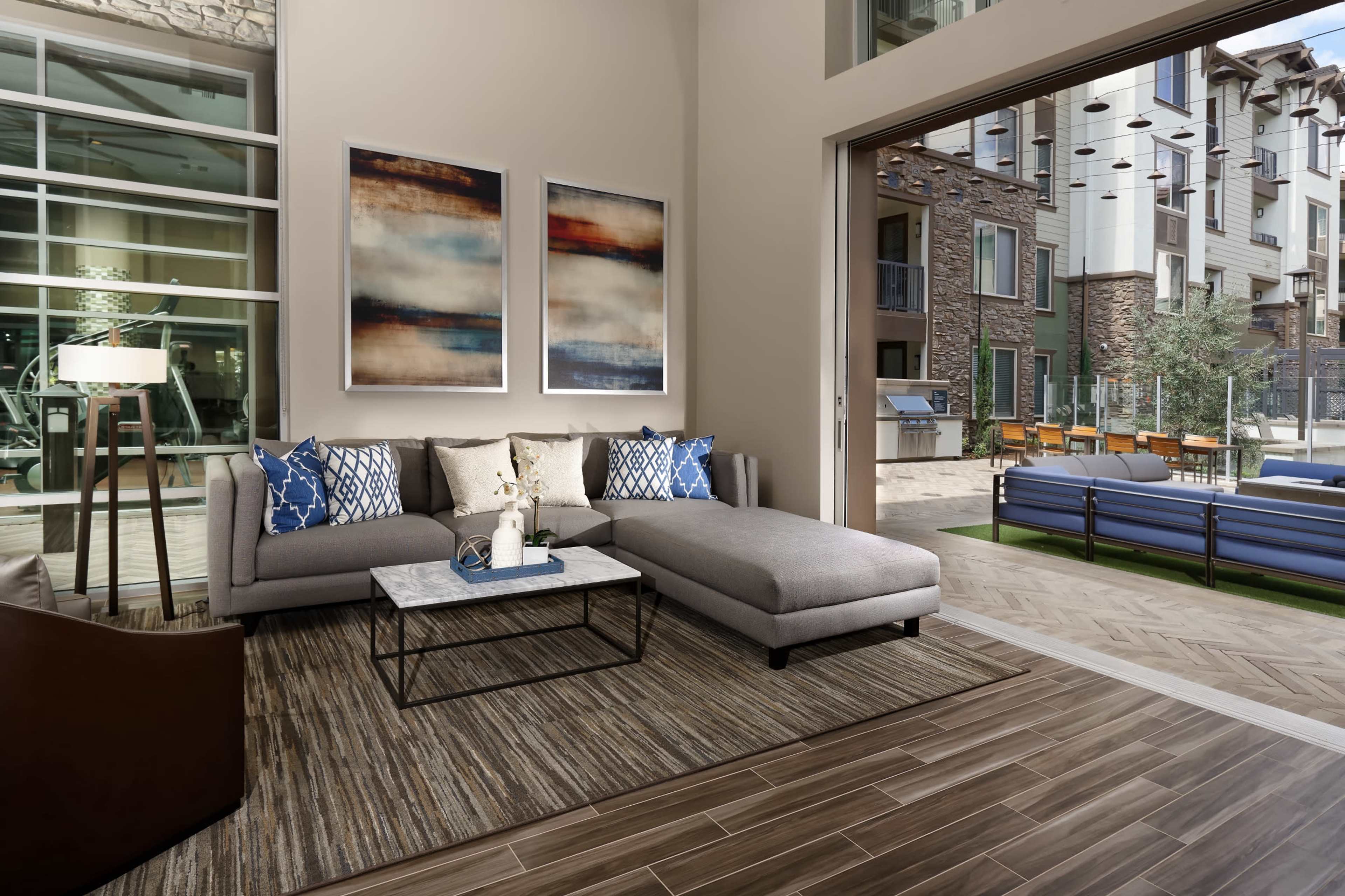 A modern living room area with a gray sectional sofa, a coffee table, and large windows overlooking a patio with seating and greenery.