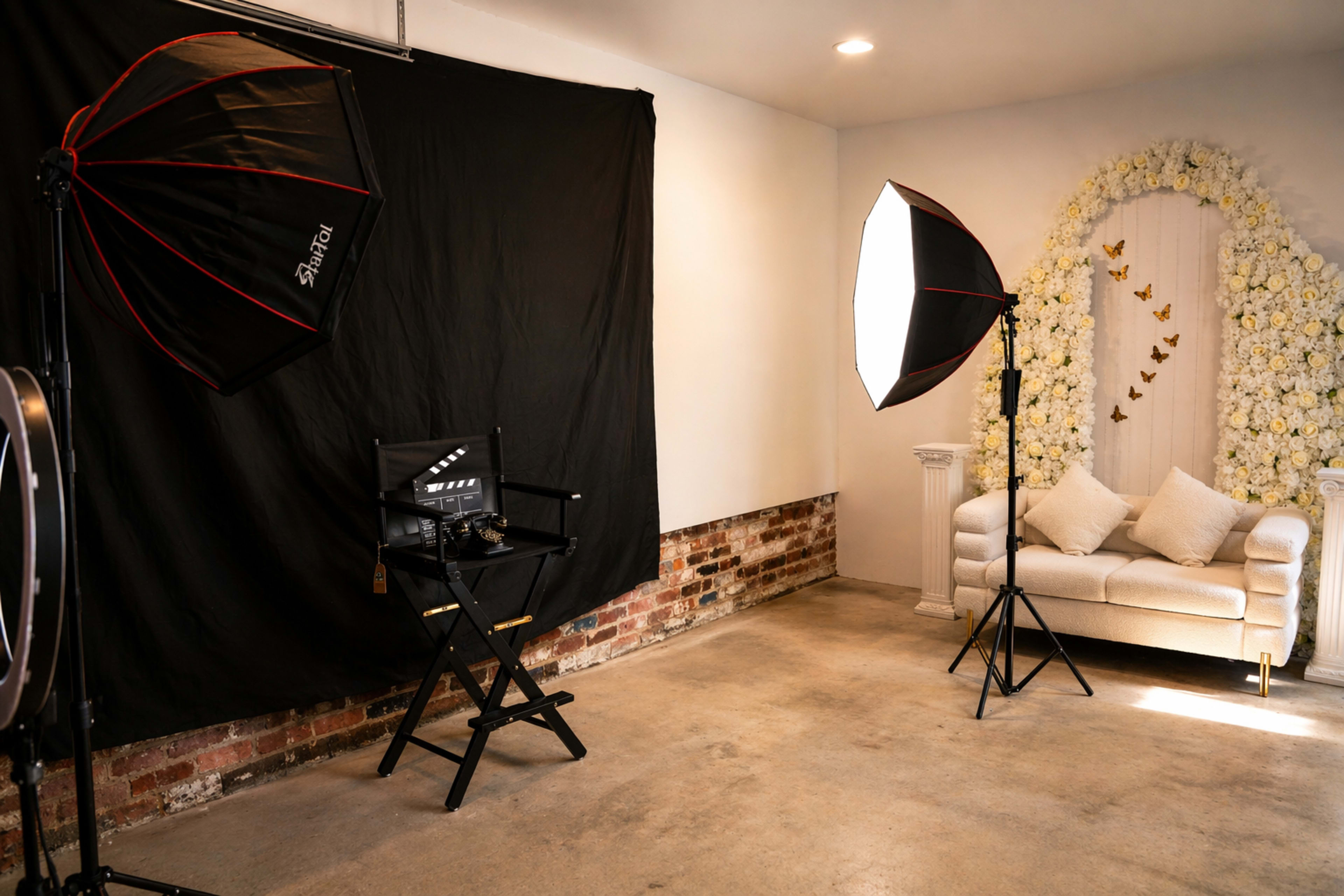 The image shows a photography studio with a black backdrop, two lighting equipment setups, a director's chair, and a white sofa adorned with floral decorations and butterflies.