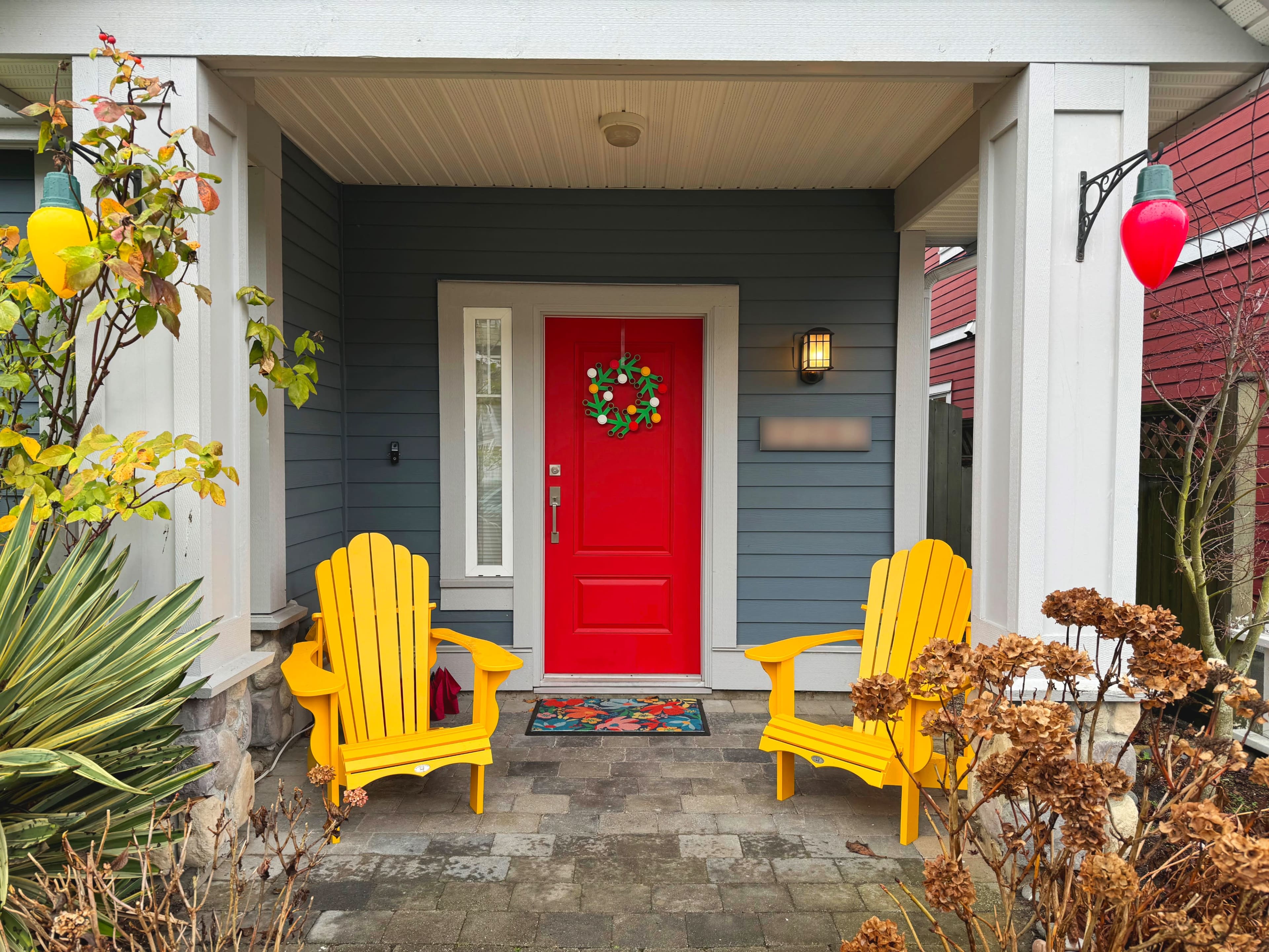 A front porch featuring two yellow Adirondack chairs, a red door with a wreath, and a stone pathway surrounded by plants.