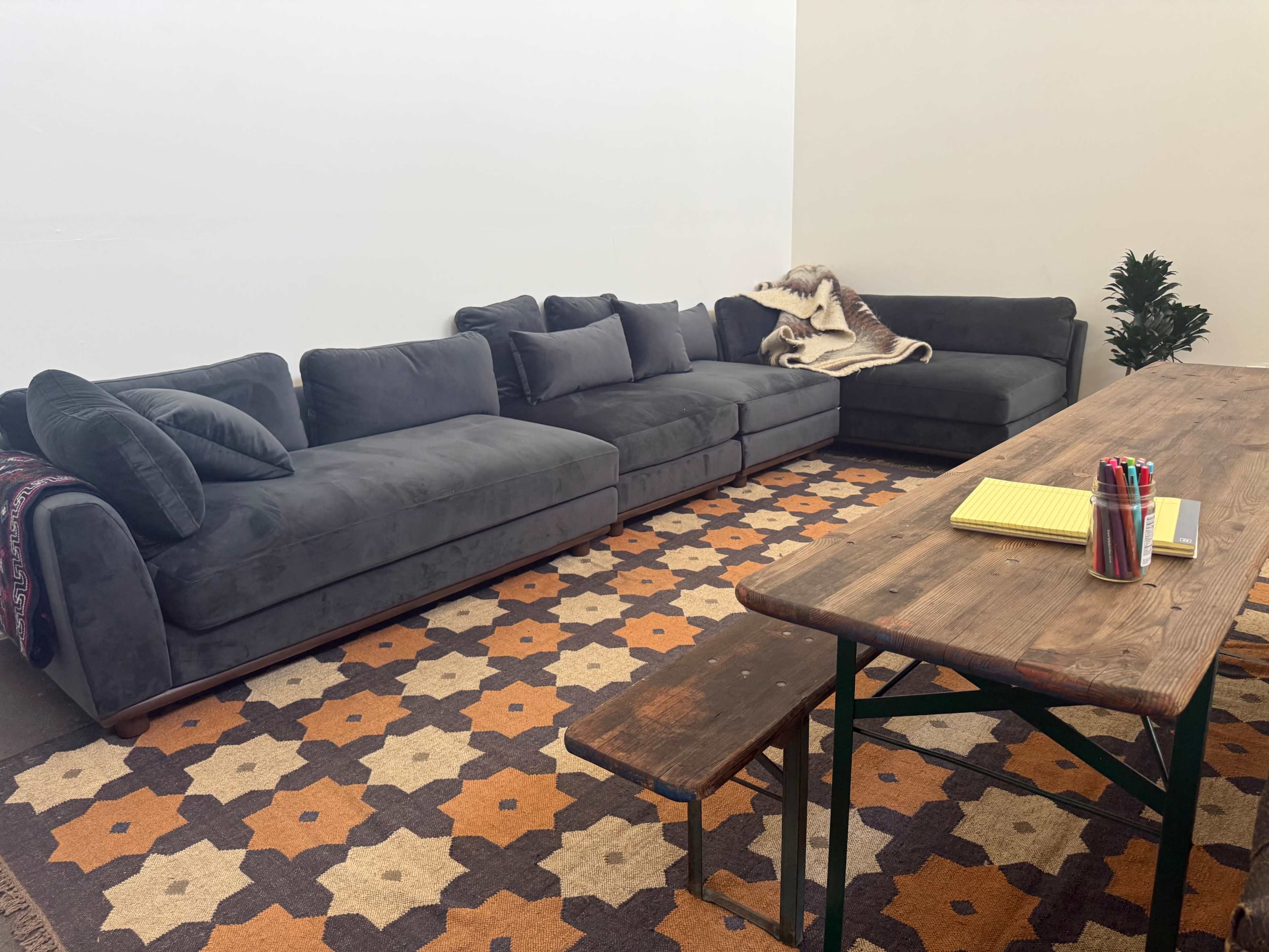 The image shows a modern living space with a large gray sectional sofa, a wooden table with stationery, and a patterned rug underneath.
