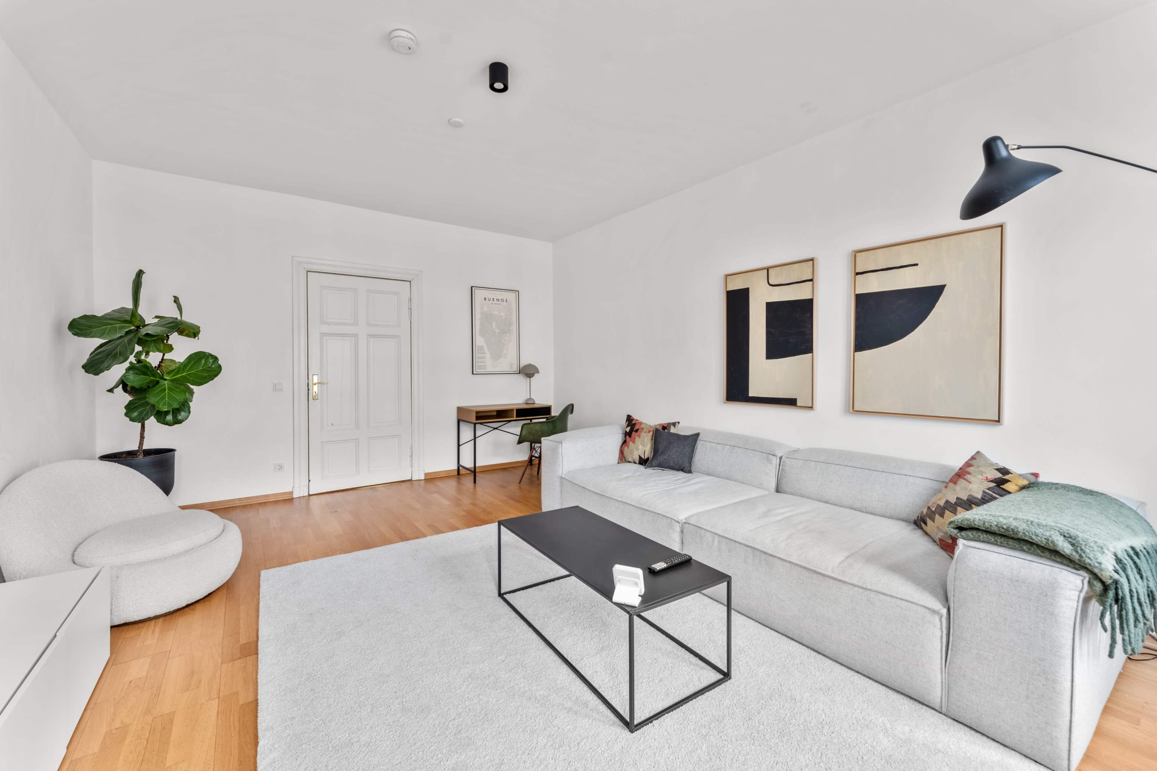 A modern living room features a gray sectional sofa, a black coffee table, two abstract wall art pieces, and a potted plant next to a door.
