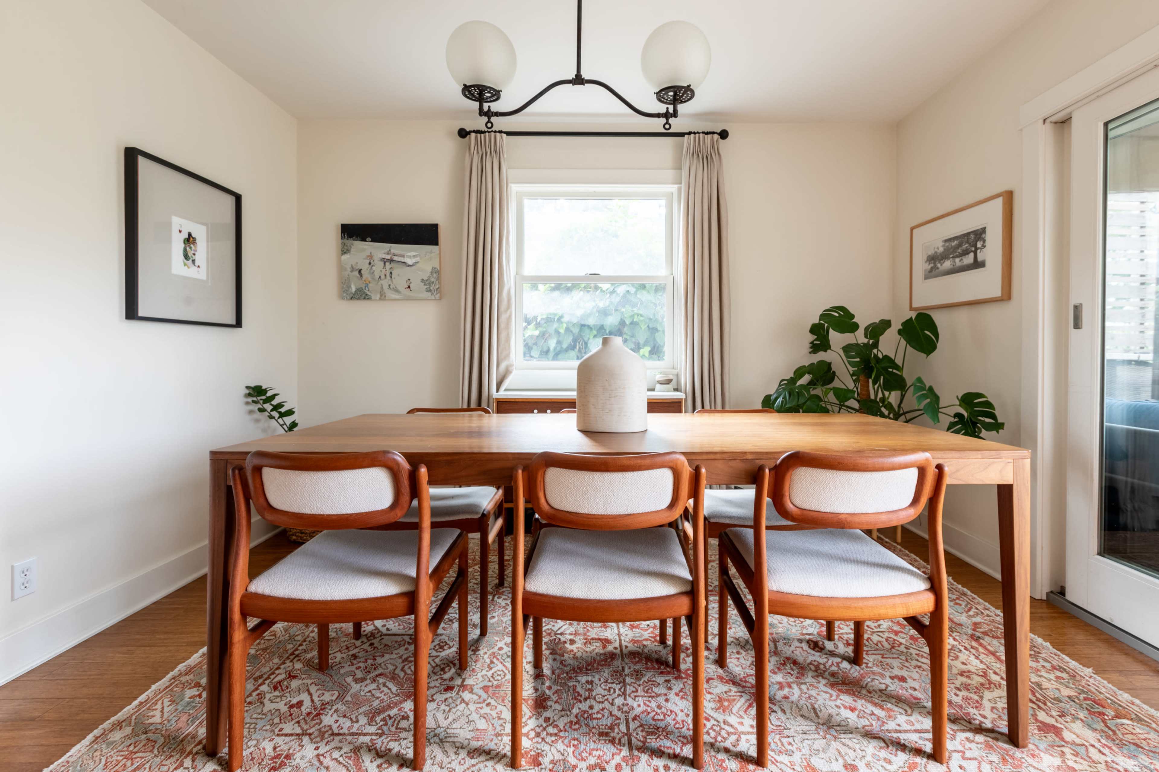 A wooden dining table accompanied by six chairs is set in a brightly lit room with large windows and decorative plants.