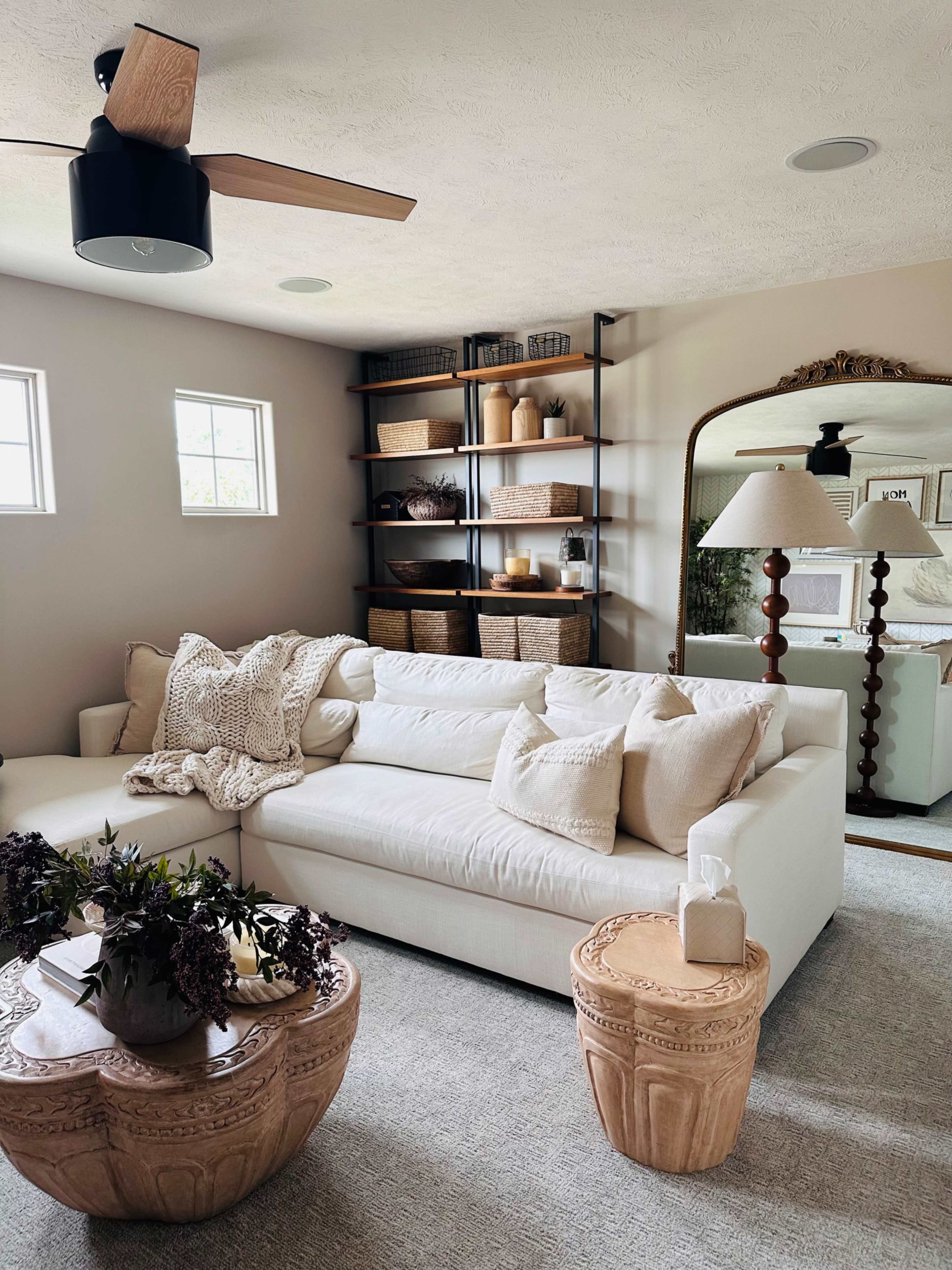 The image shows a modern living room featuring a white sofa with decorative pillows, a wooden coffee table, and a decorative mirror on the wall.