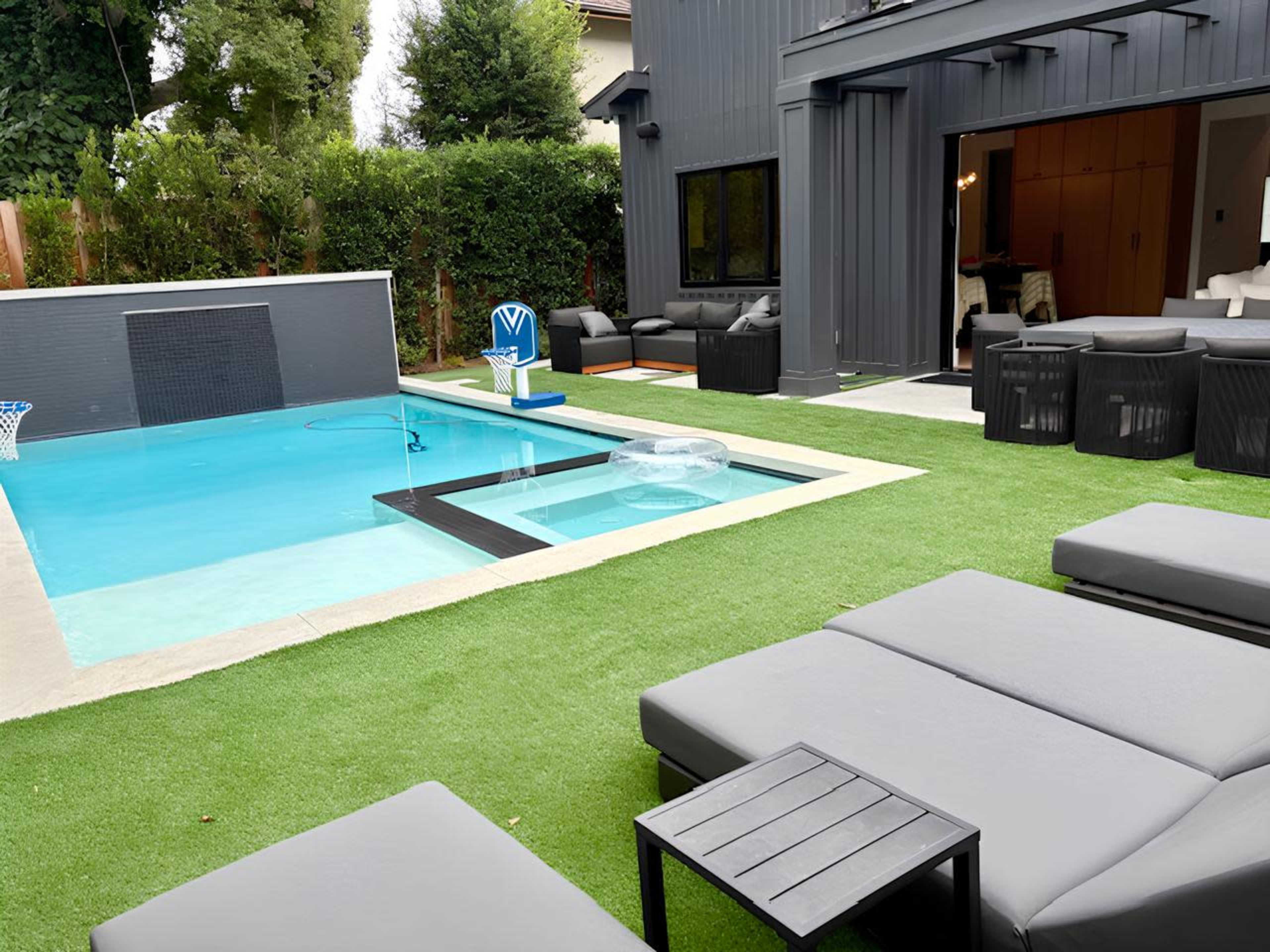 The image shows a modern backyard featuring a swimming pool surrounded by artificial grass, outdoor furniture, and a basketball hoop.