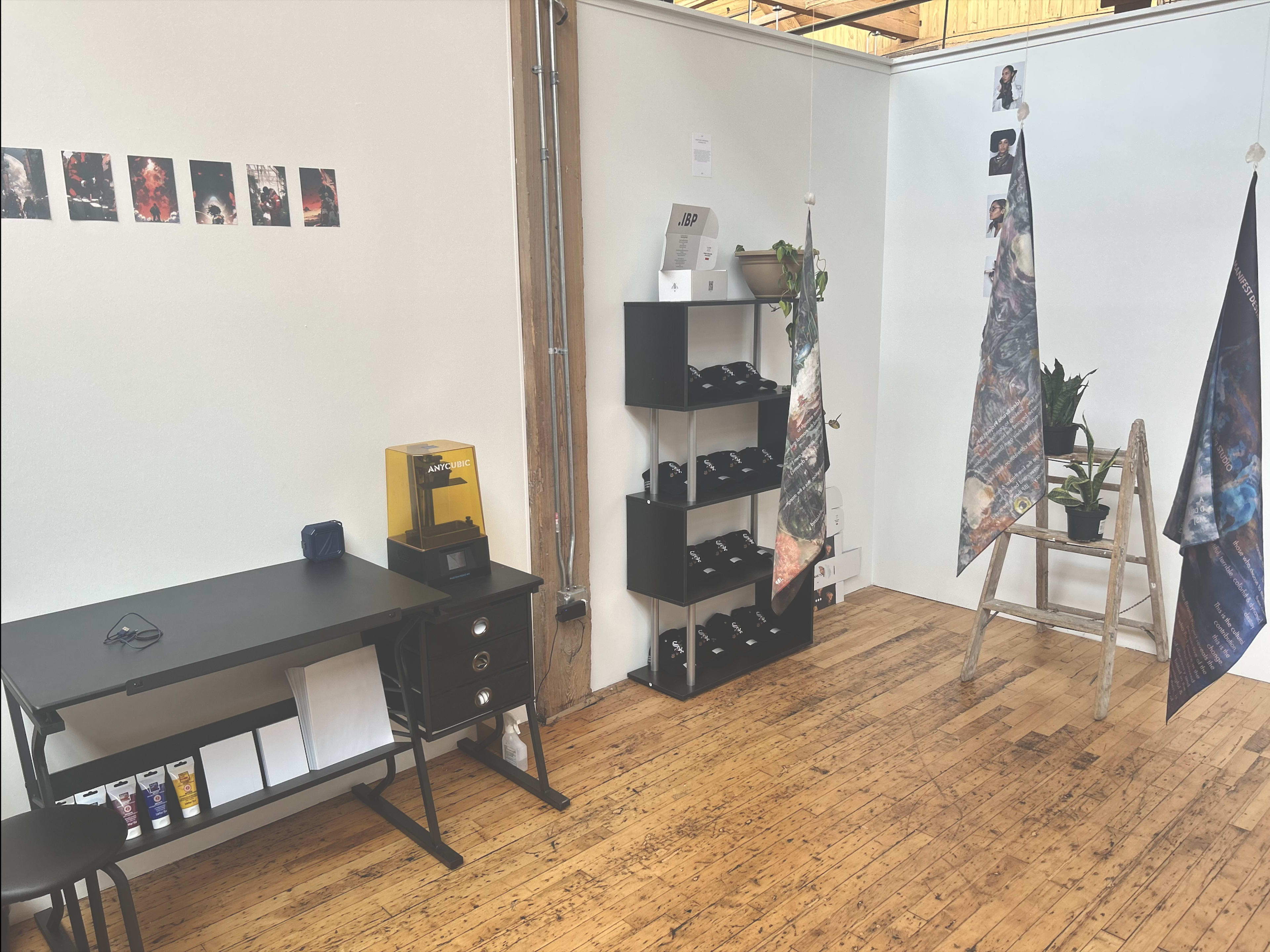 A minimalist exhibition space featuring a black desk, a shelf with various items, and two vertical banners hanging, set against white walls and wooden flooring.