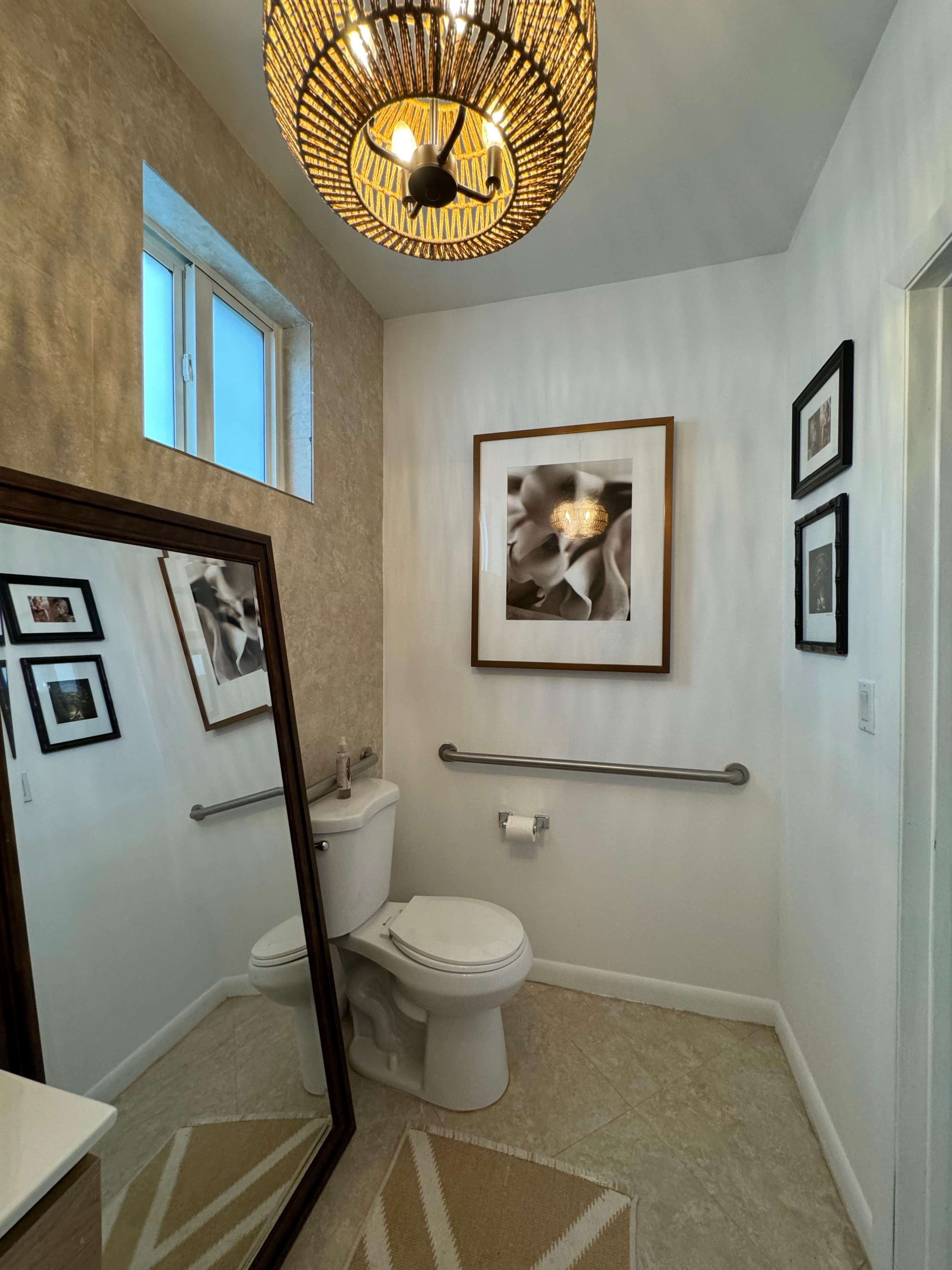 A small bathroom features a toilet, a large mirror, a decorative pendant light, and framed photographs on the walls.