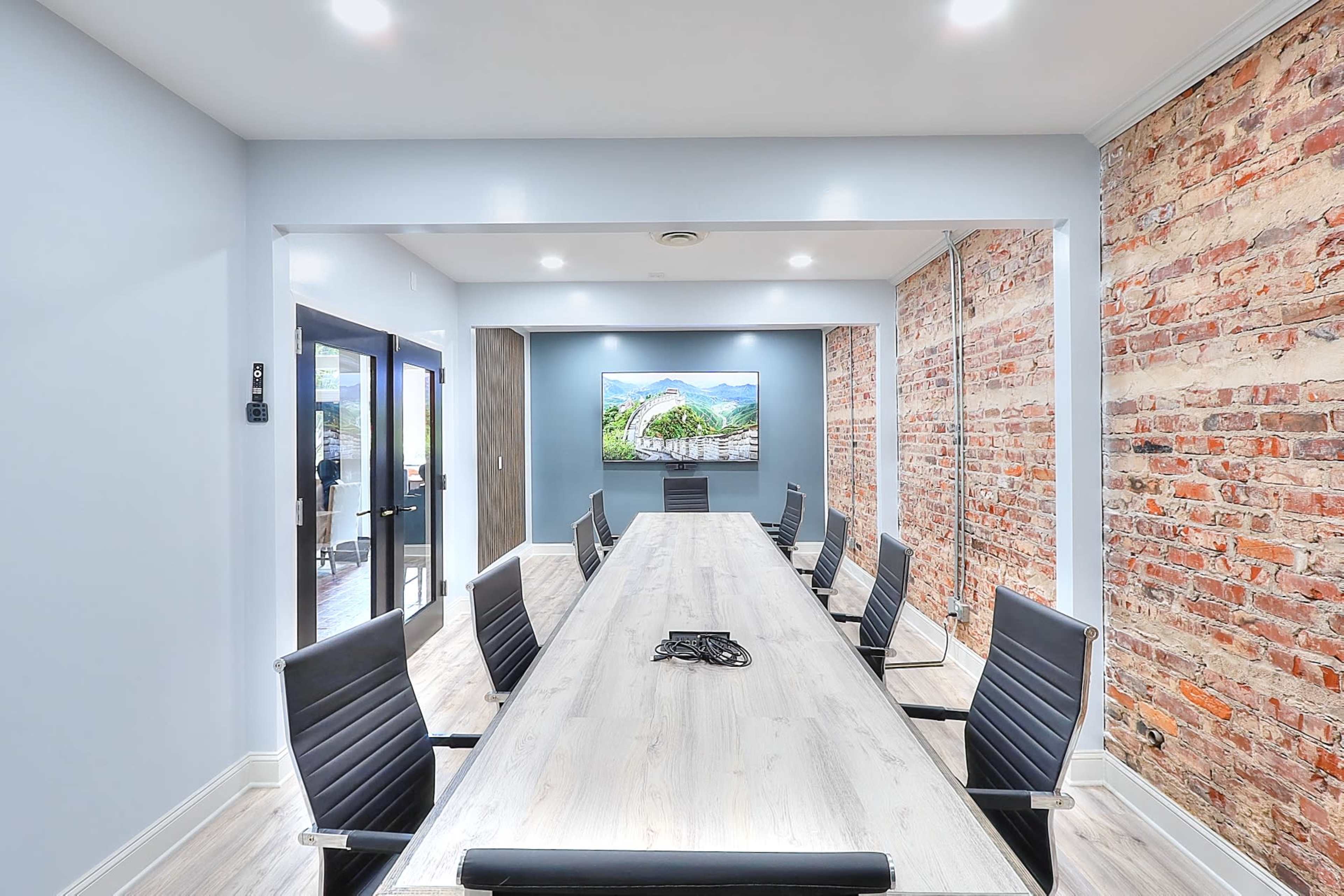 A modern conference room features a long wooden table surrounded by black chairs, with exposed brick walls and a large screen displaying a landscape.