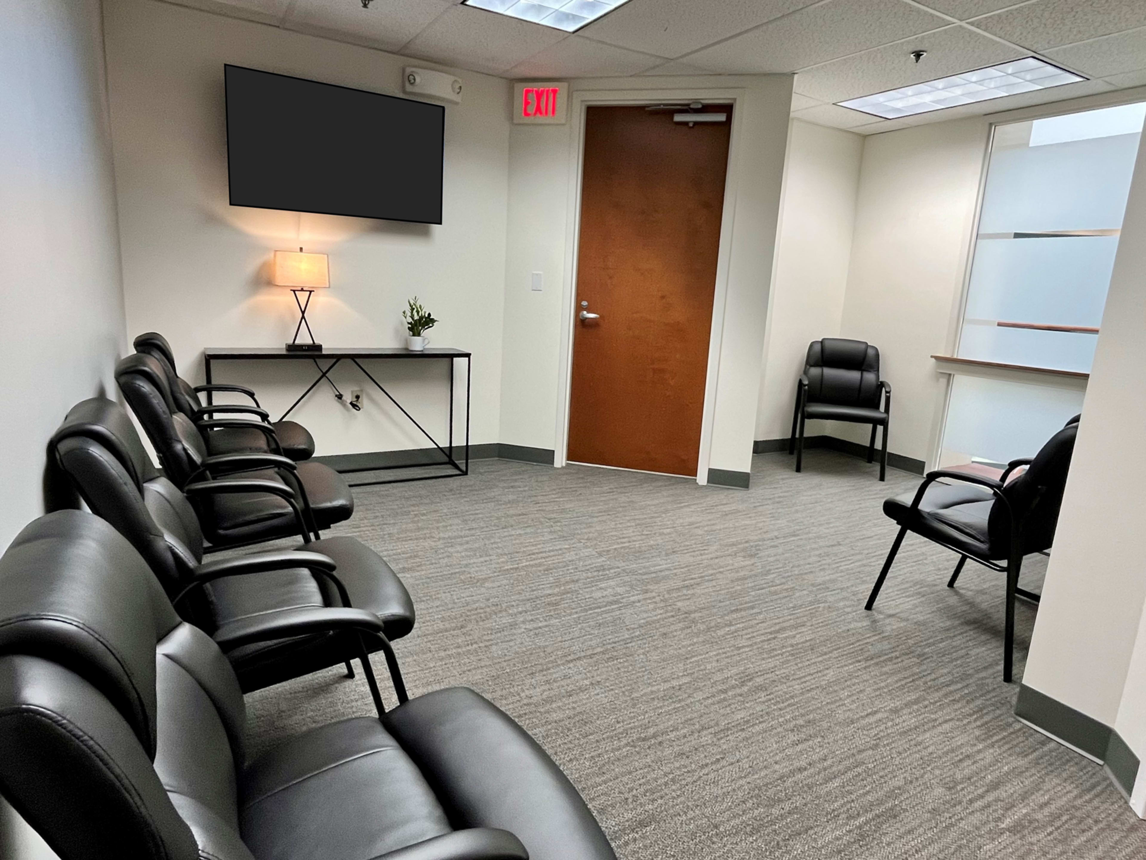 The image shows a waiting room with several black chairs arranged along the wall, a small table with a lamp, a wall-mounted screen, and a door leading out of the room.