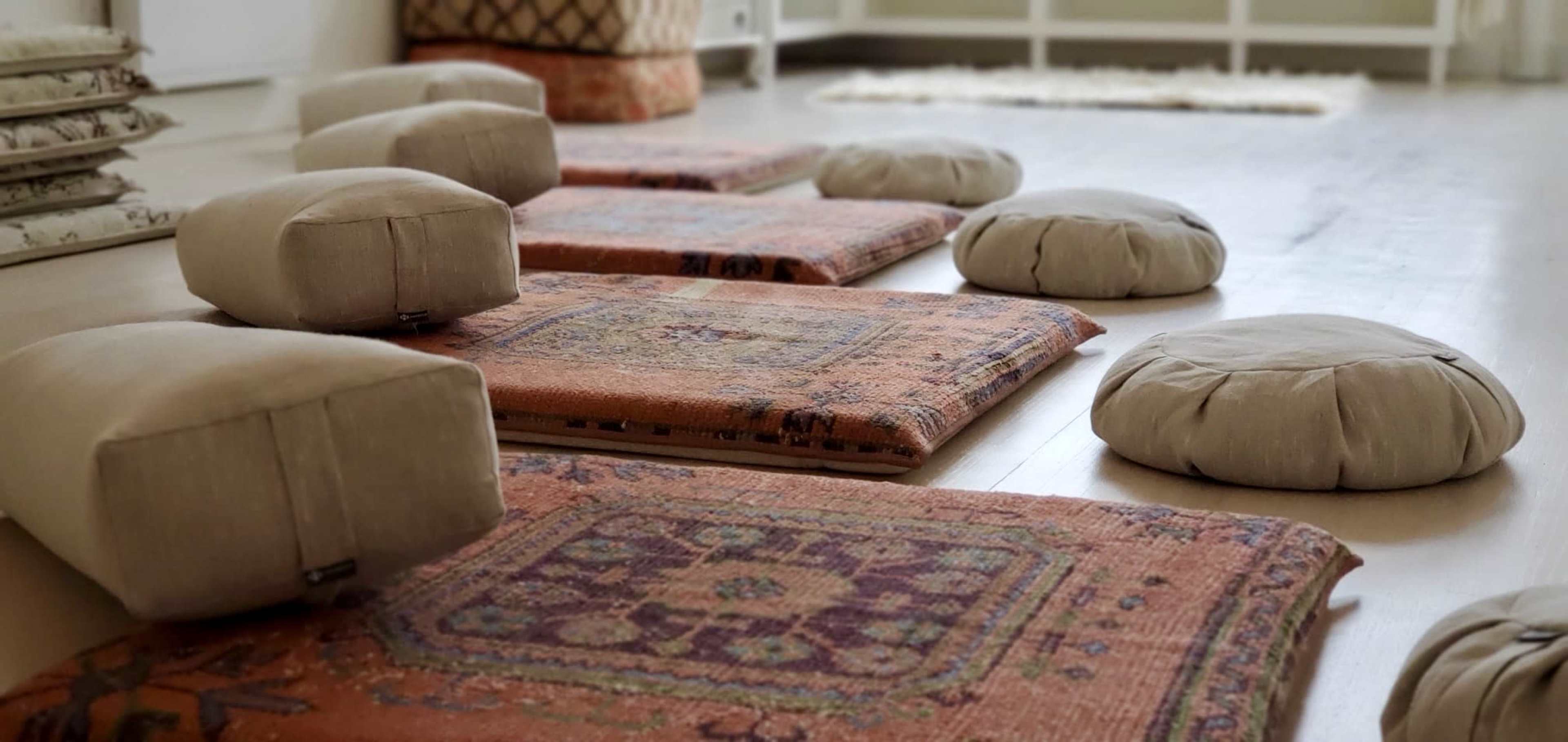 The image shows a serene indoor space with several cushions and intricately patterned rugs laid out on a wooden floor.