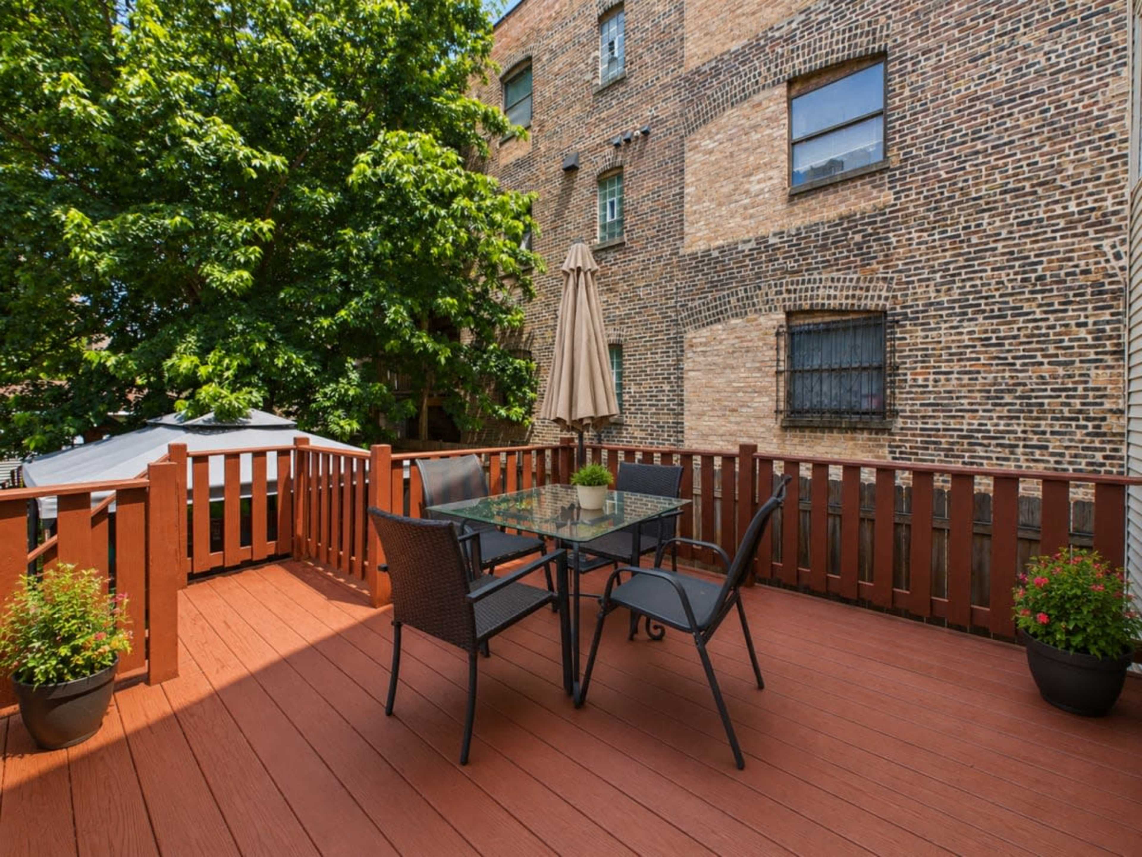A wooden deck with a glass table and chairs is surrounded by greenery and brick buildings.