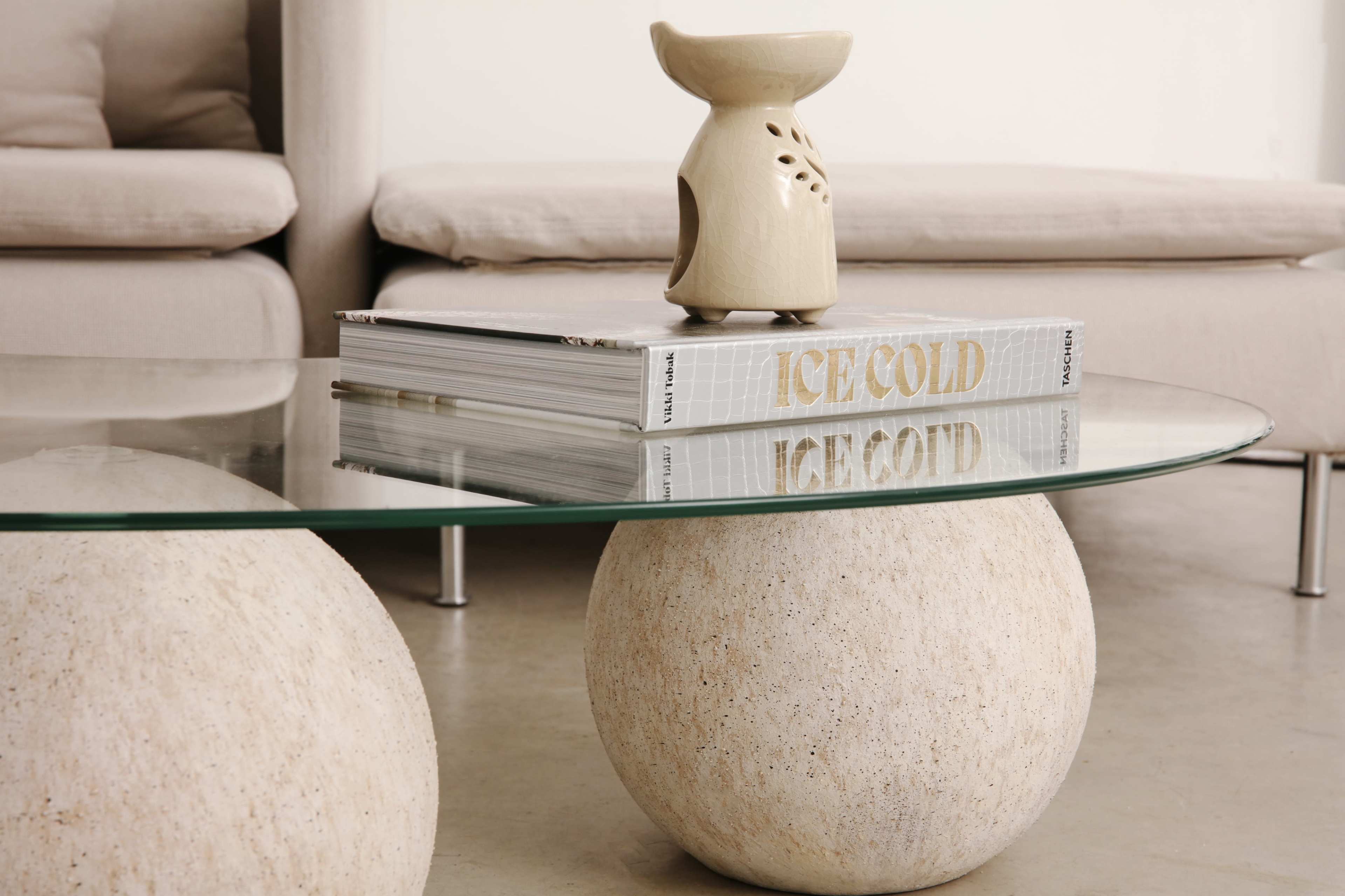 A glass coffee table with stone spheres as supports holds a magazine titled "ICE COLD" and a decorative ceramic oil burner.