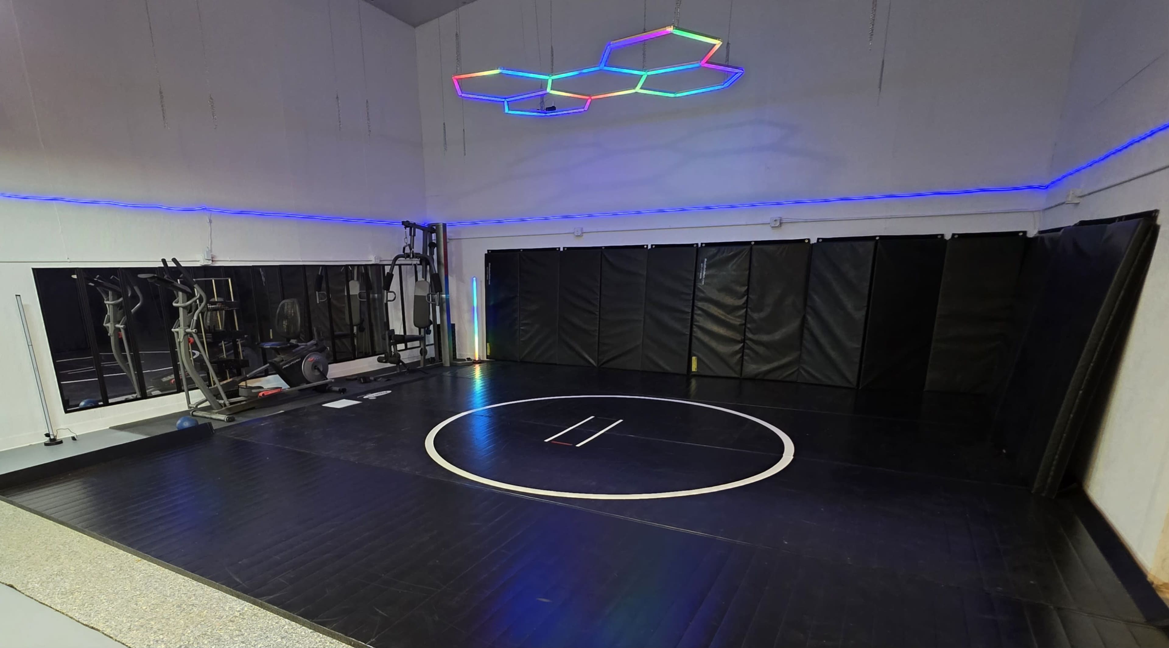 The image shows a wrestling gym with a circular mat in the center, surrounded by padded walls and various gym equipment along one side.