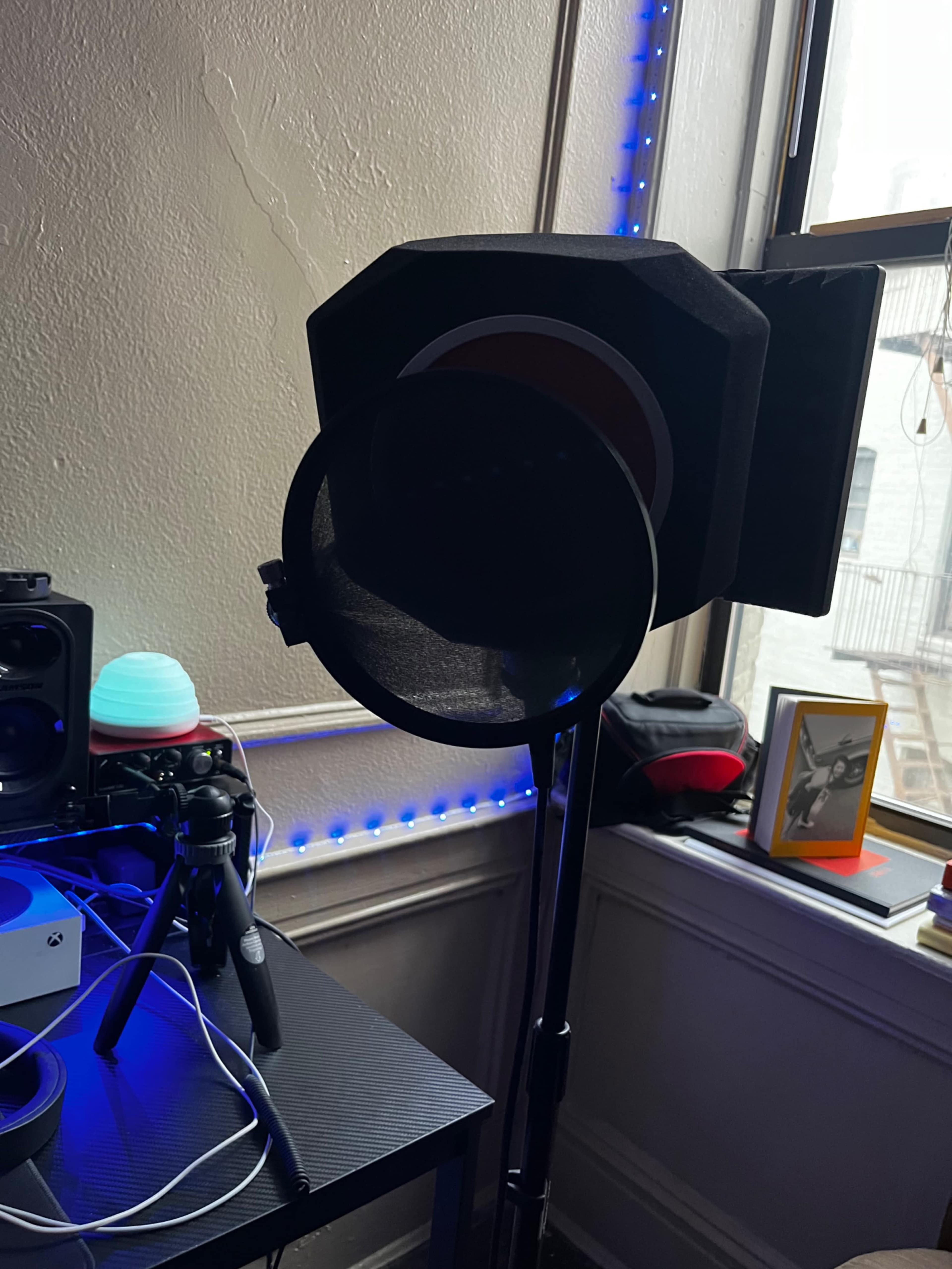 A microphone setup with a pop filter stands beside a colorful speaker and various audio equipment in a sunlit room.
