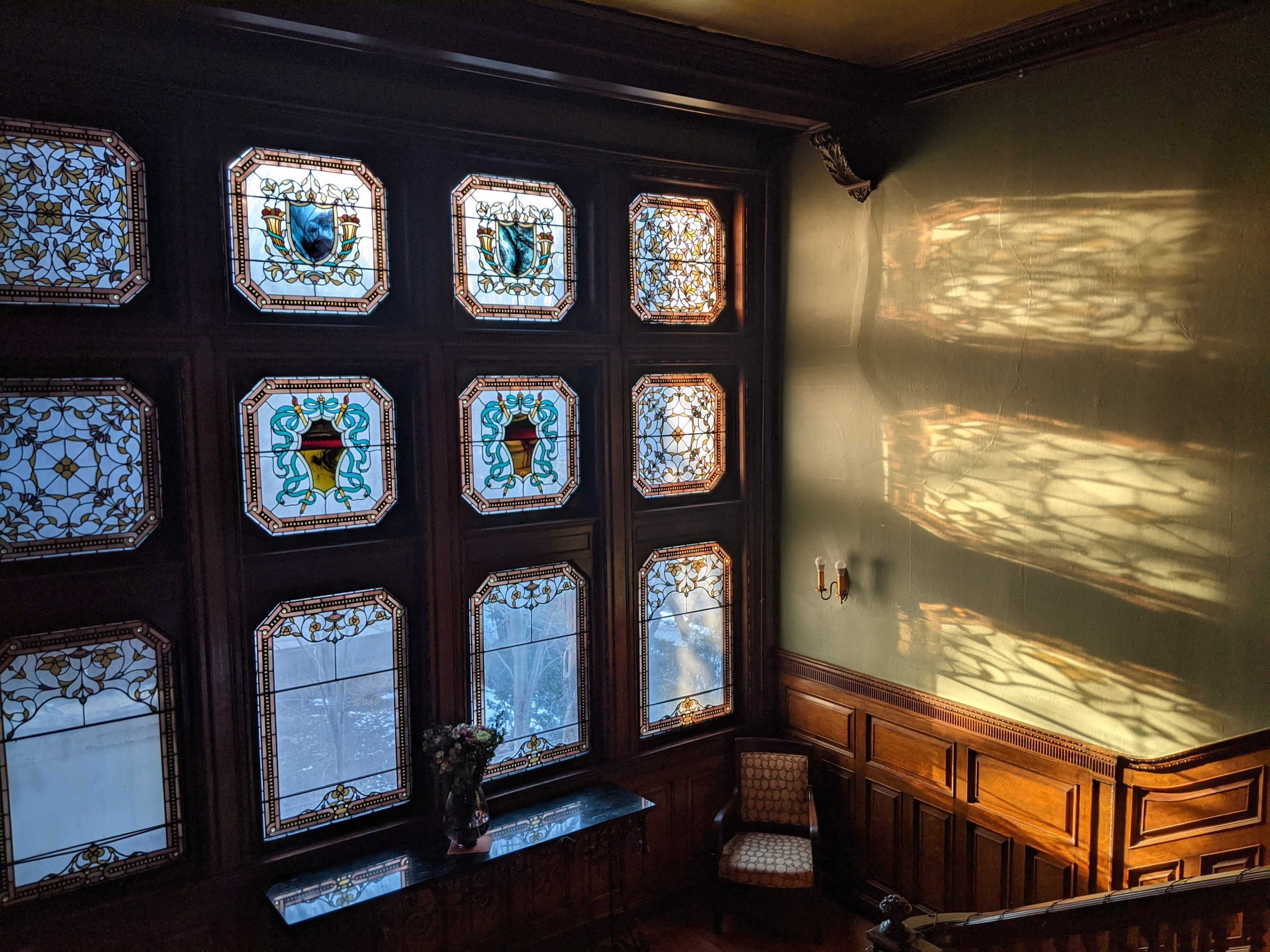 A staircase area with large stained glass windows casting colorful shadows on the wall and wooden paneling.