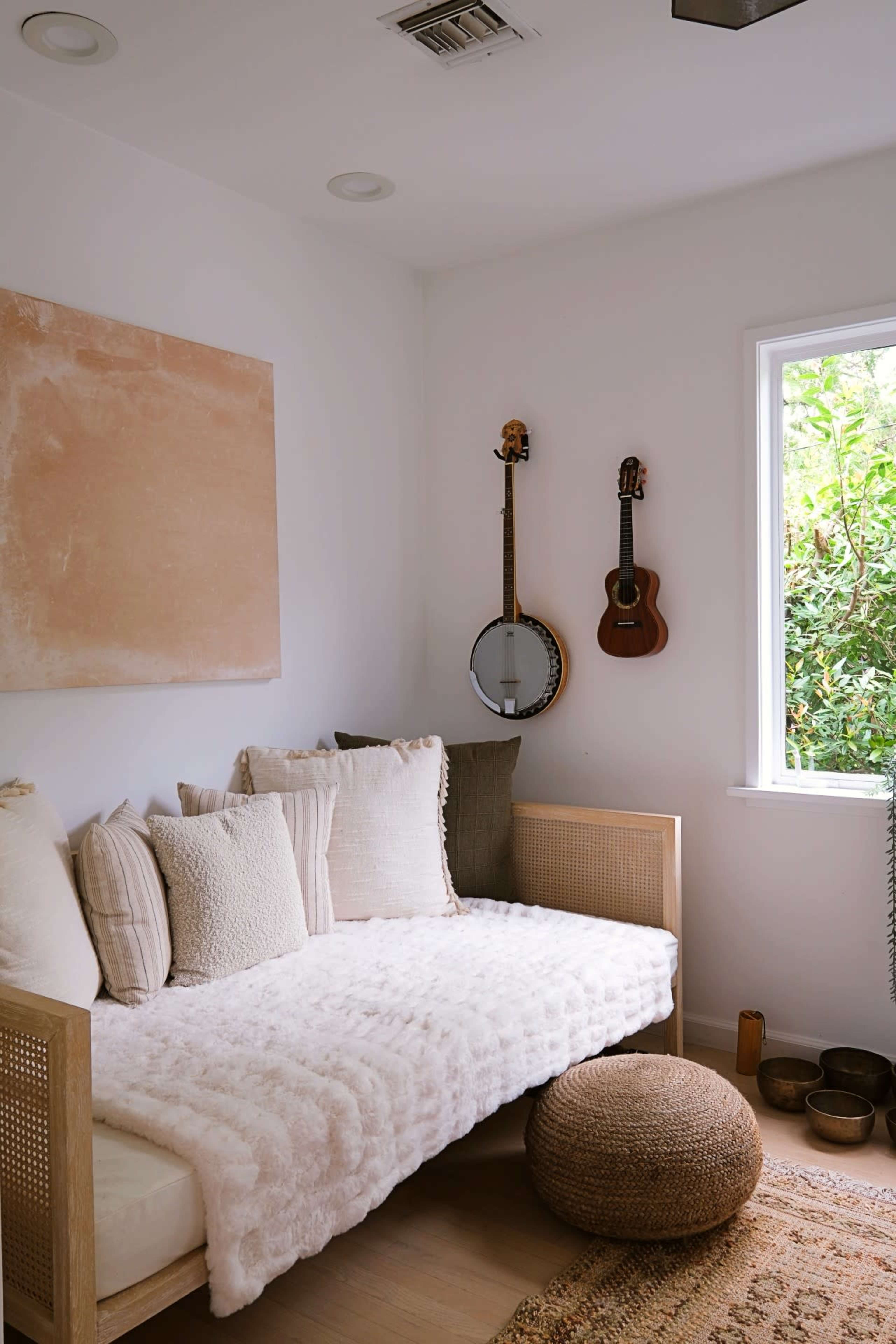 A cozy room with a daybed covered in a light blanket, flanked by decorative pillows, and featuring two hanging musical instruments beside a window.