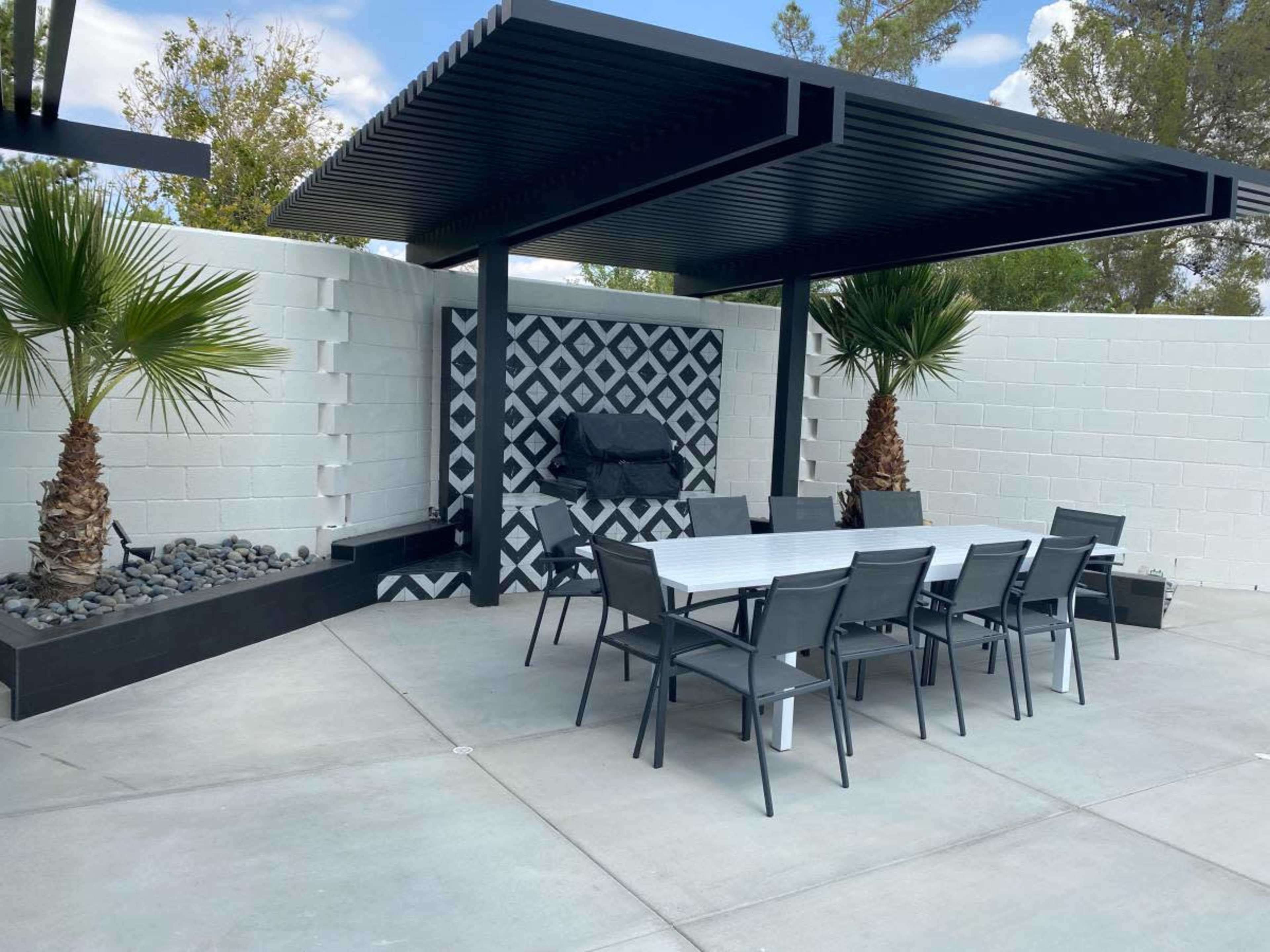 A modern outdoor dining area features a long table and chairs beneath a black pergola, with a patterned accent wall and palm trees nearby.