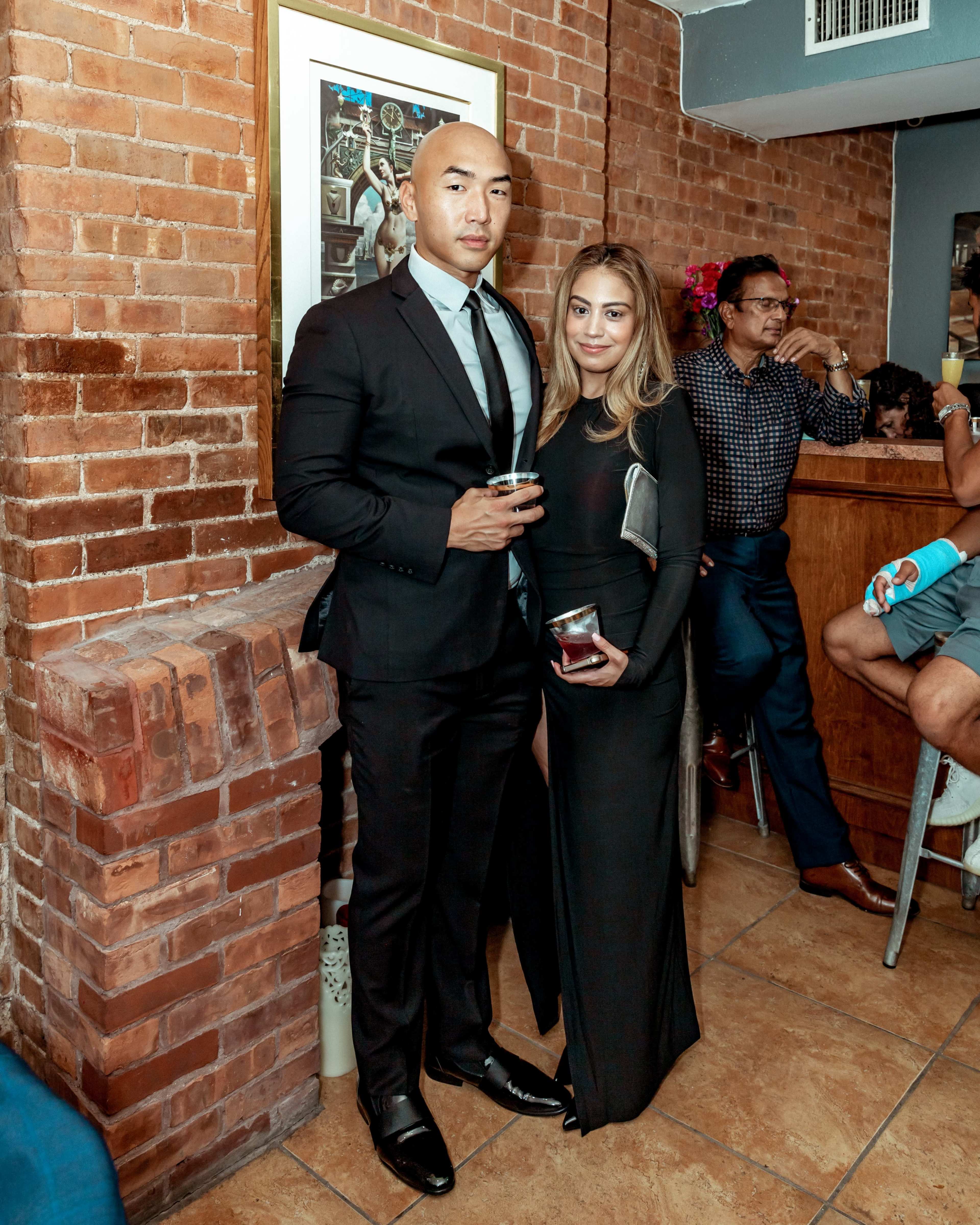 A man in a black suit stands next to a woman in a black dress, both holding drinks, in a brick-walled bar with other patrons in the background.