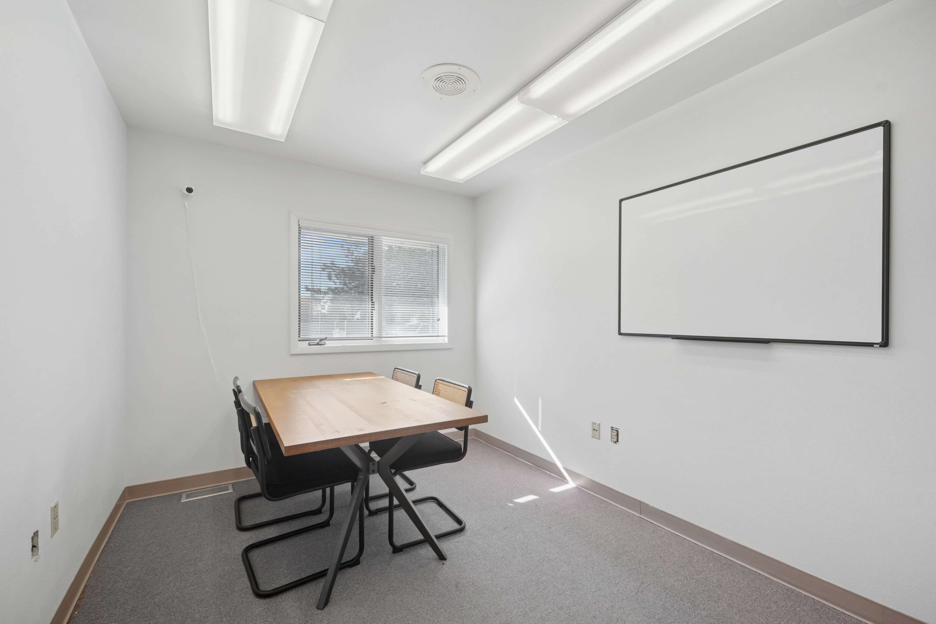 A small meeting room features a wooden table with four black chairs, a whiteboard on the wall, and a window allowing natural light.