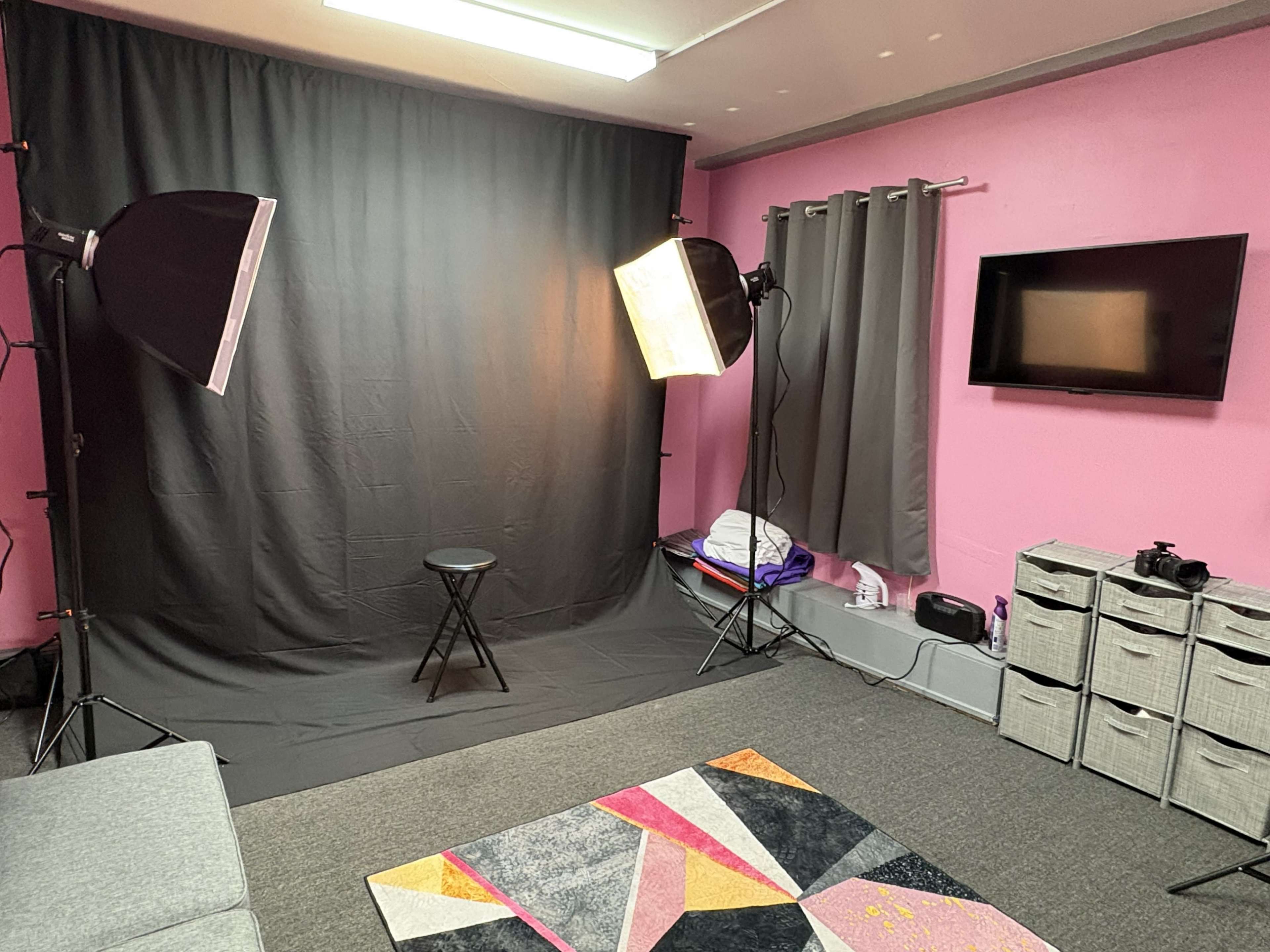 The image shows a photography studio setup featuring a black backdrop, two softbox lights, a stool in the center, a colorful rug, and a television mounted on a pink wall.