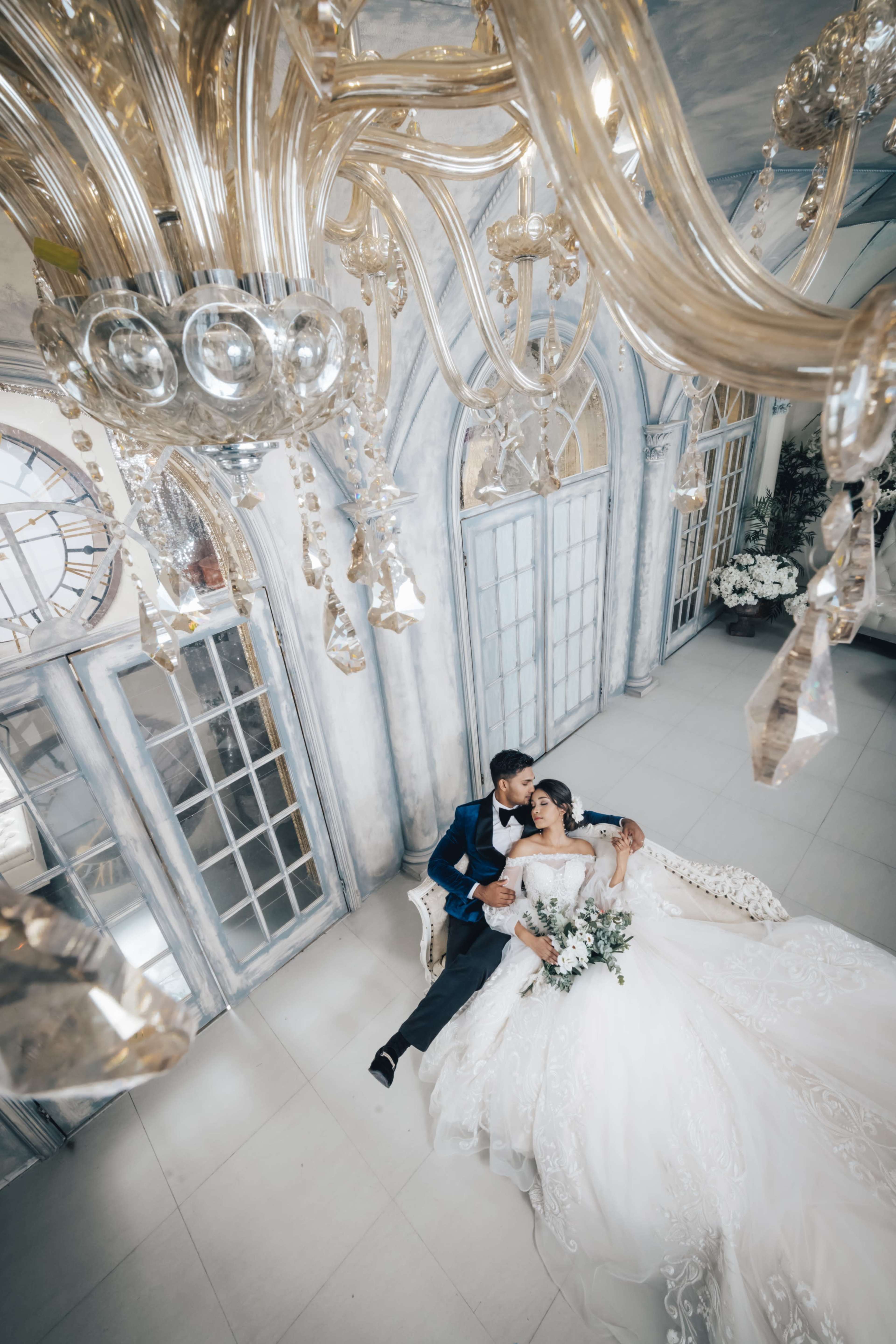 A bride and groom sit together on a couch beneath an ornate chandelier in a beautifully decorated interior.