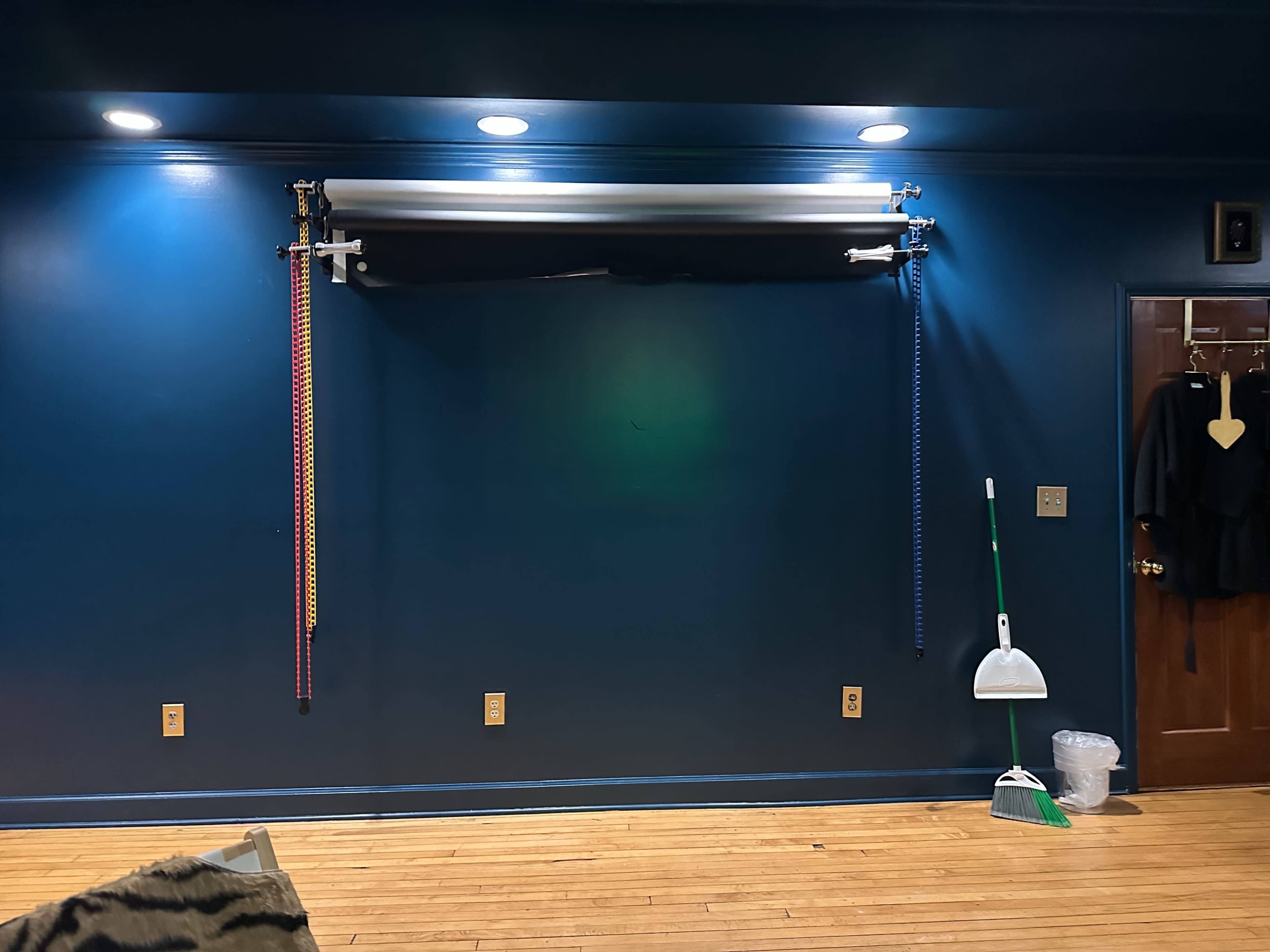 The image shows a dark blue wall with two colorful straps hanging from a mounted rod, accompanied by a broom and dustpan in the corner.